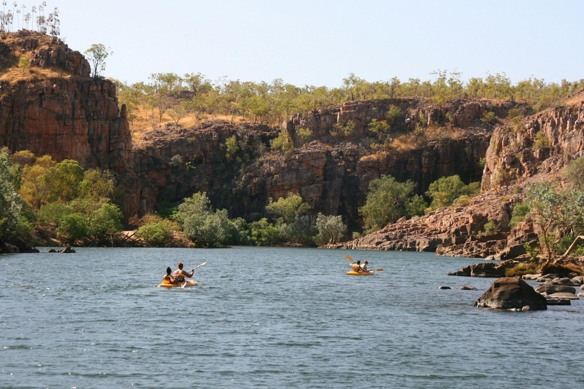 Four people paddle along in small canoes in a water-filled gorge on a sunny day