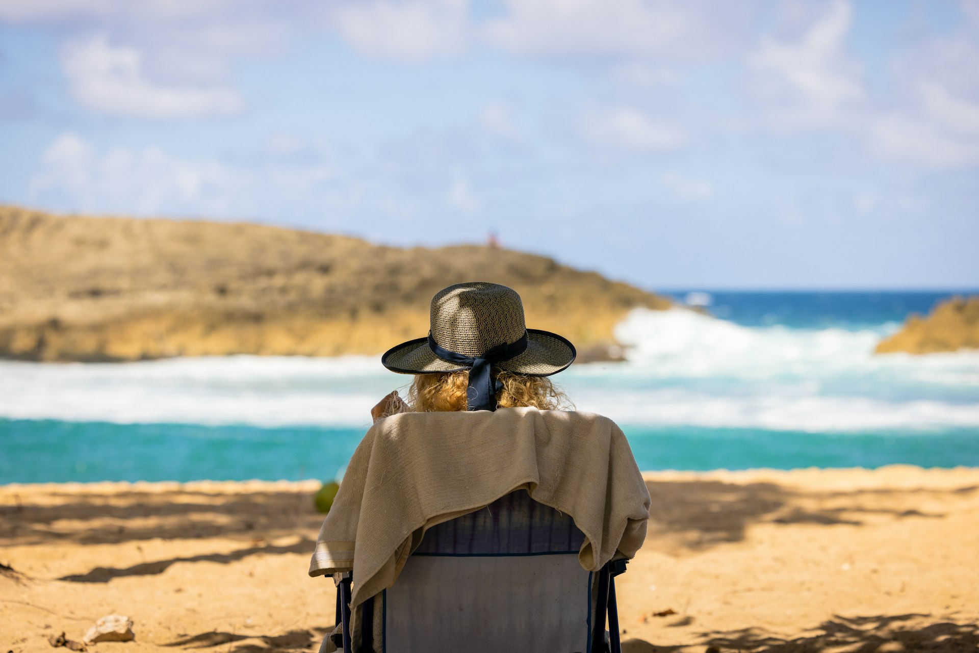 Back view of a woman sat in a chair on the beach, in the shade, with a sun hat on