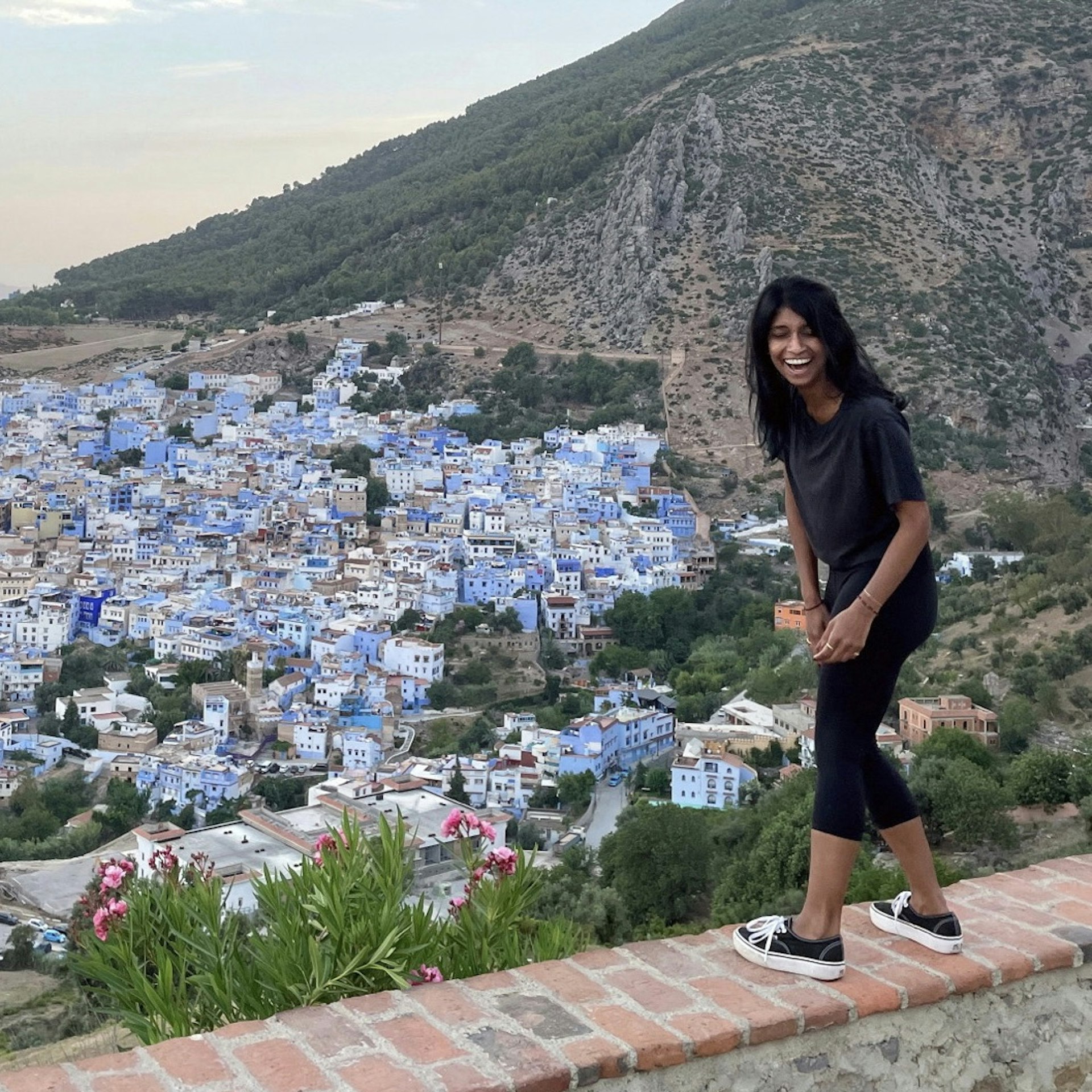 Deepa Lakshmin in Morocco, laughing and standing on a wall with the city of Chefchaouen in the background.