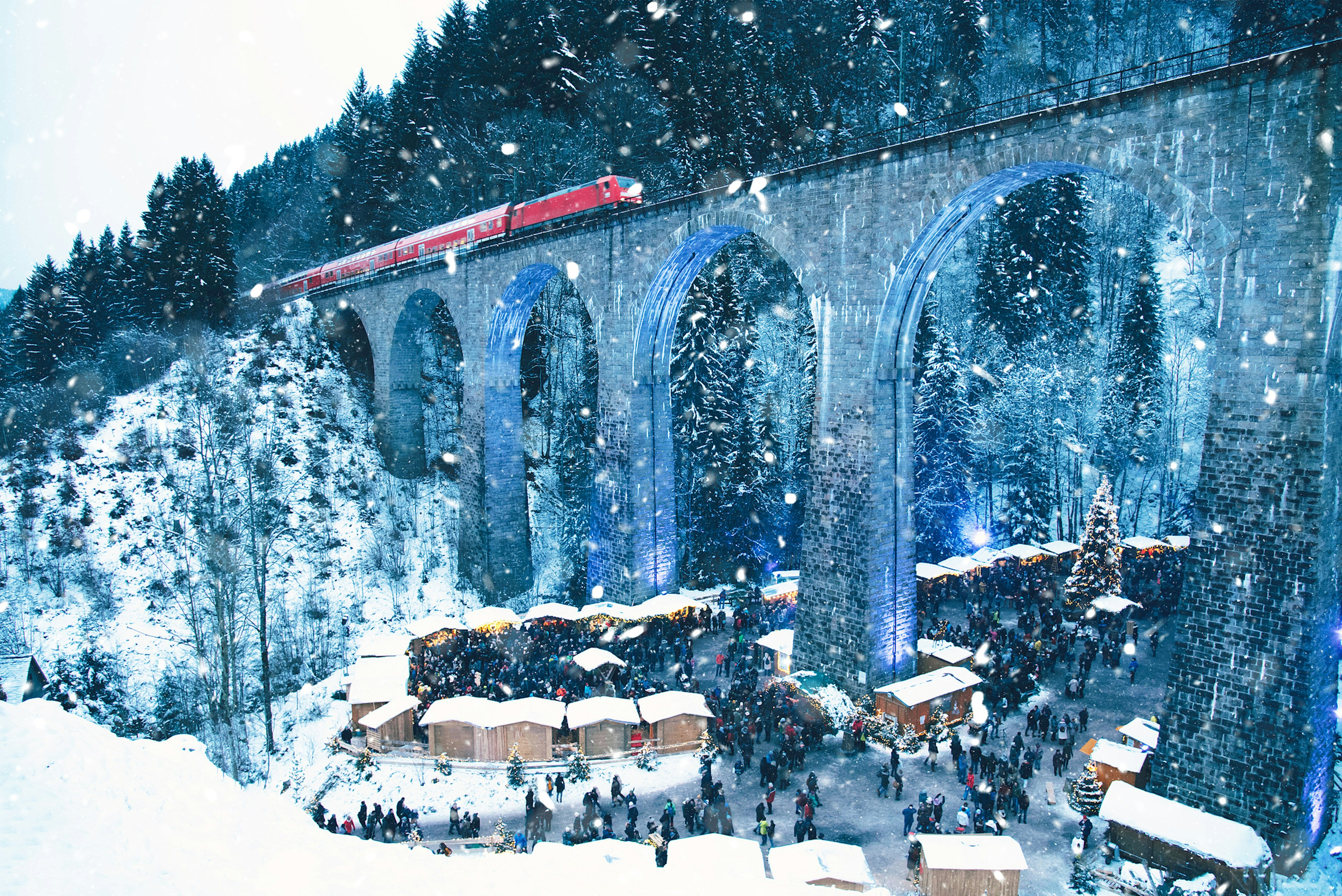 Traditional Christmas market with many wooden huts in the snow. A red train passes on a large bridge overhead