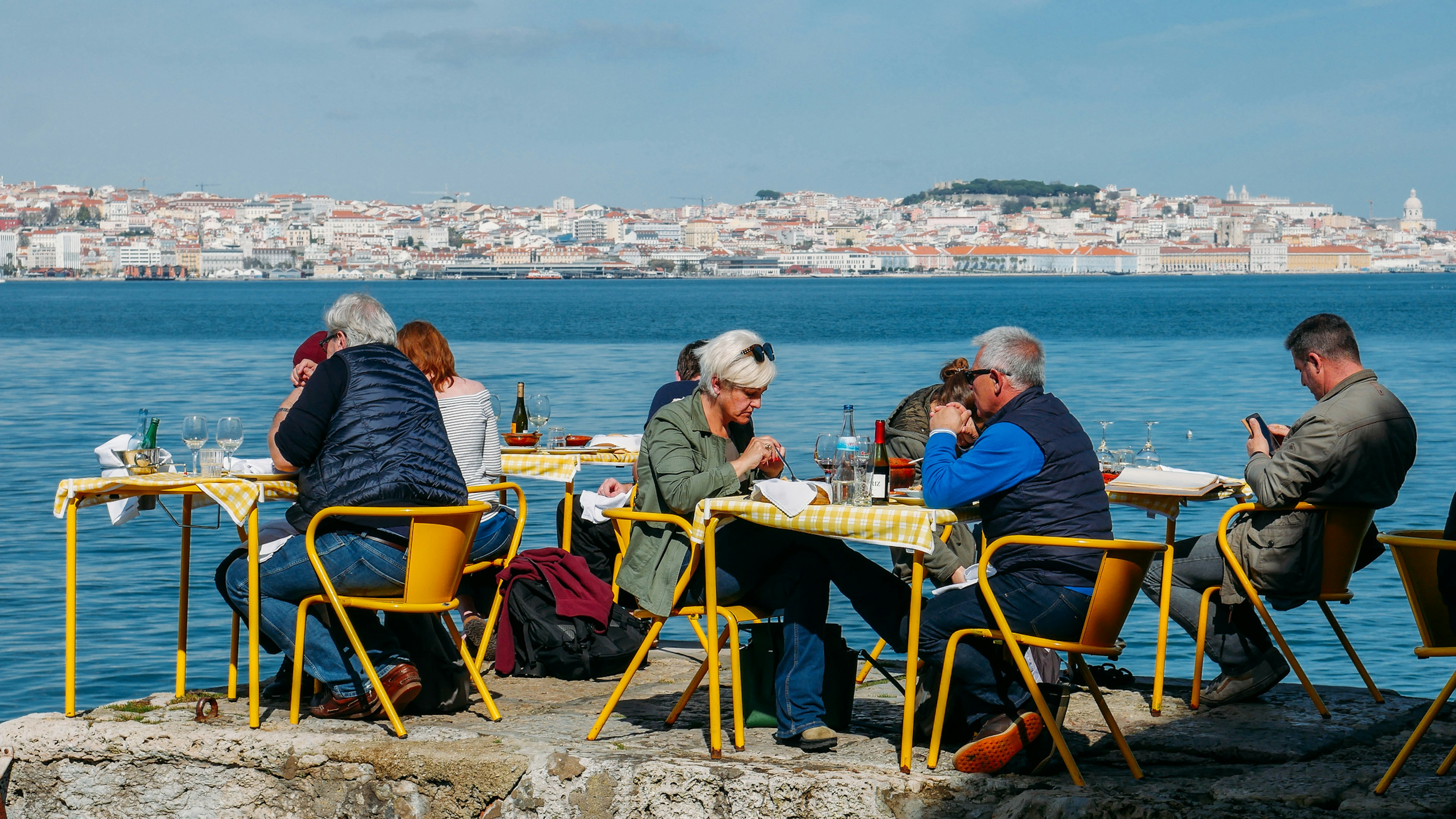 People sitting at yellow restaurant tables in the shore of the river Tagus in Cacilhas - Lisbon's cityscape in the background