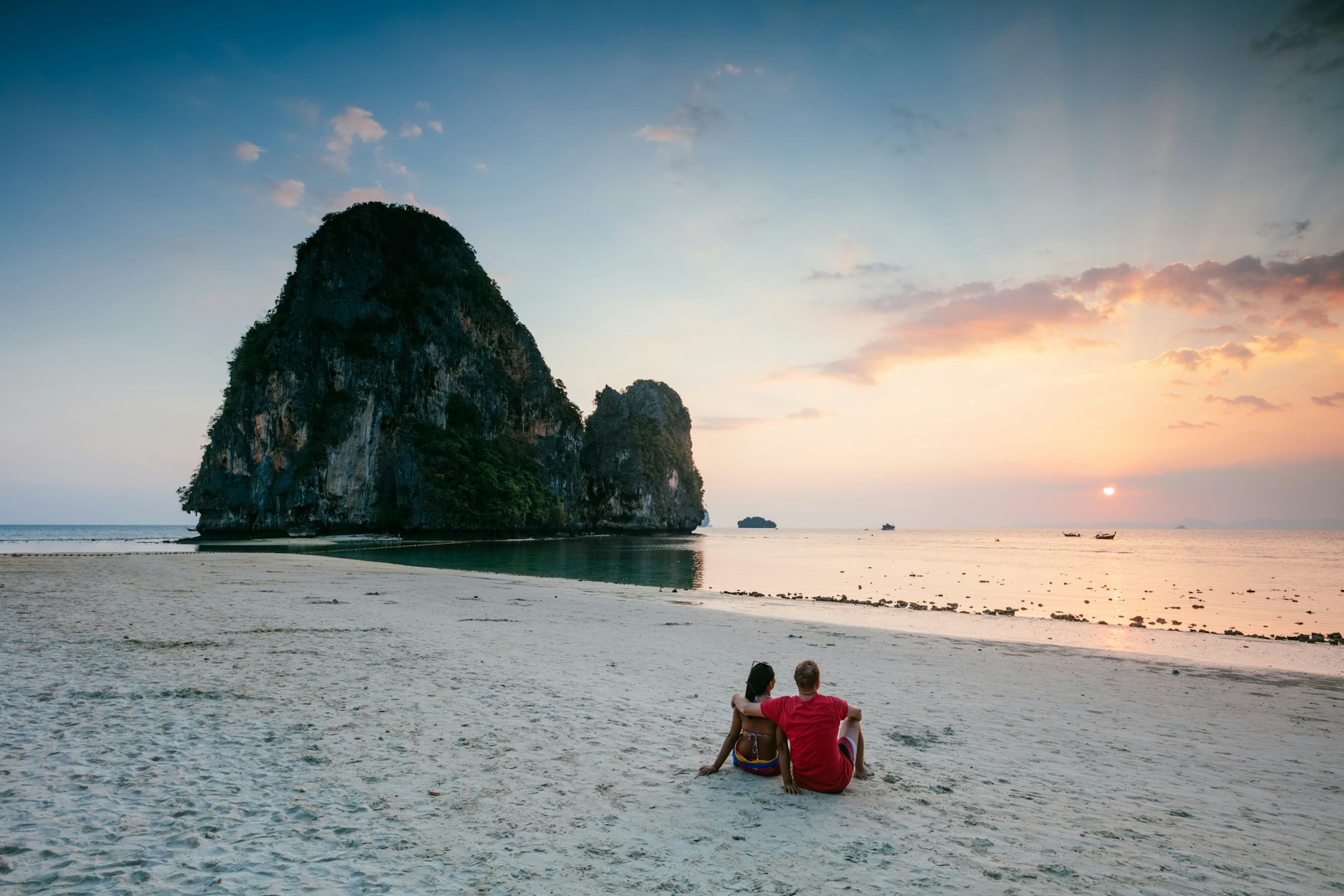 Two people sit together on a sandy beach staring out to sea where the sun sets behind a huge rocky outcrop