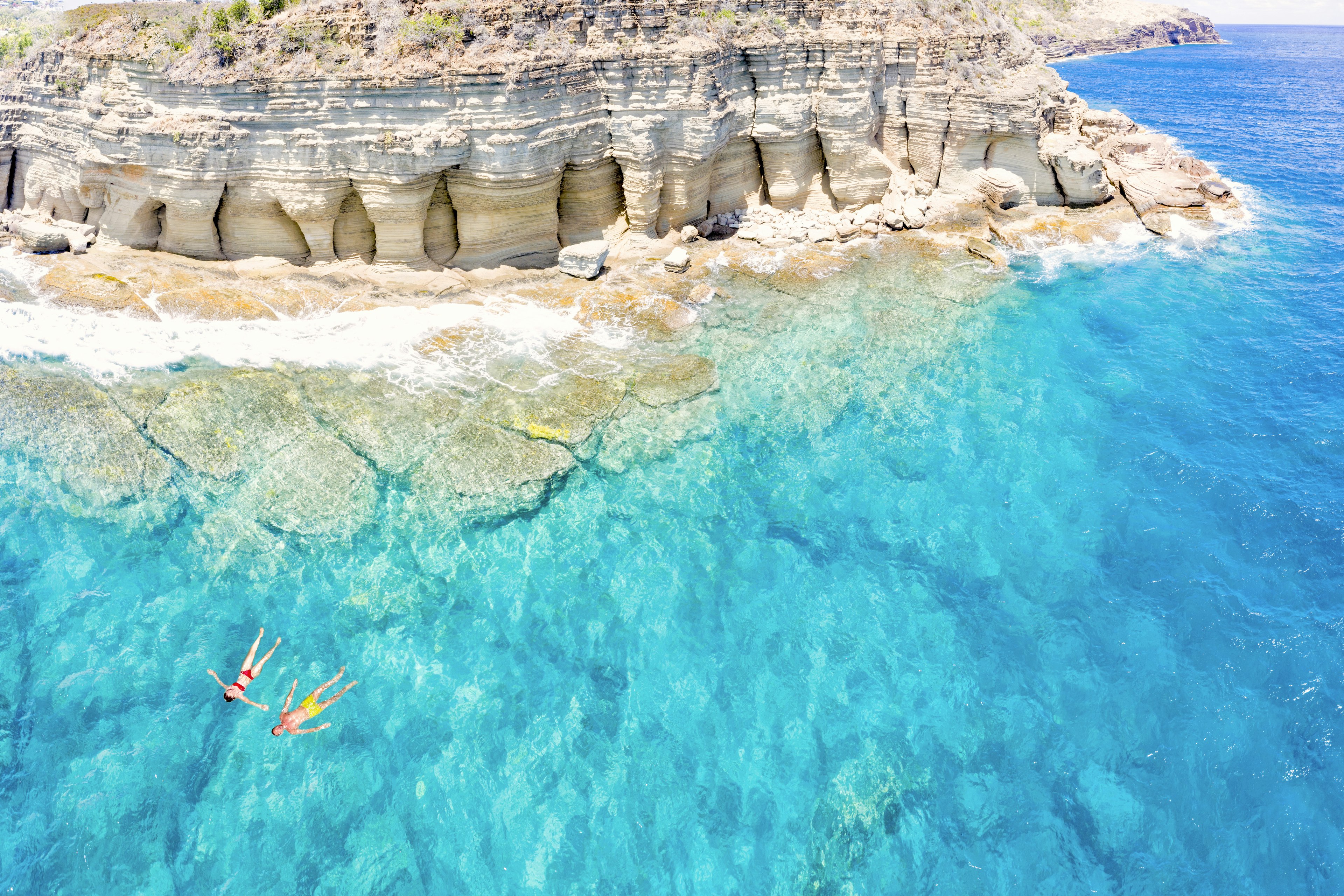 Aerial panoramic of man and woman floating in the turquoise sea near Pillar of Hercules, English Harbour, Antigua, Caribbean