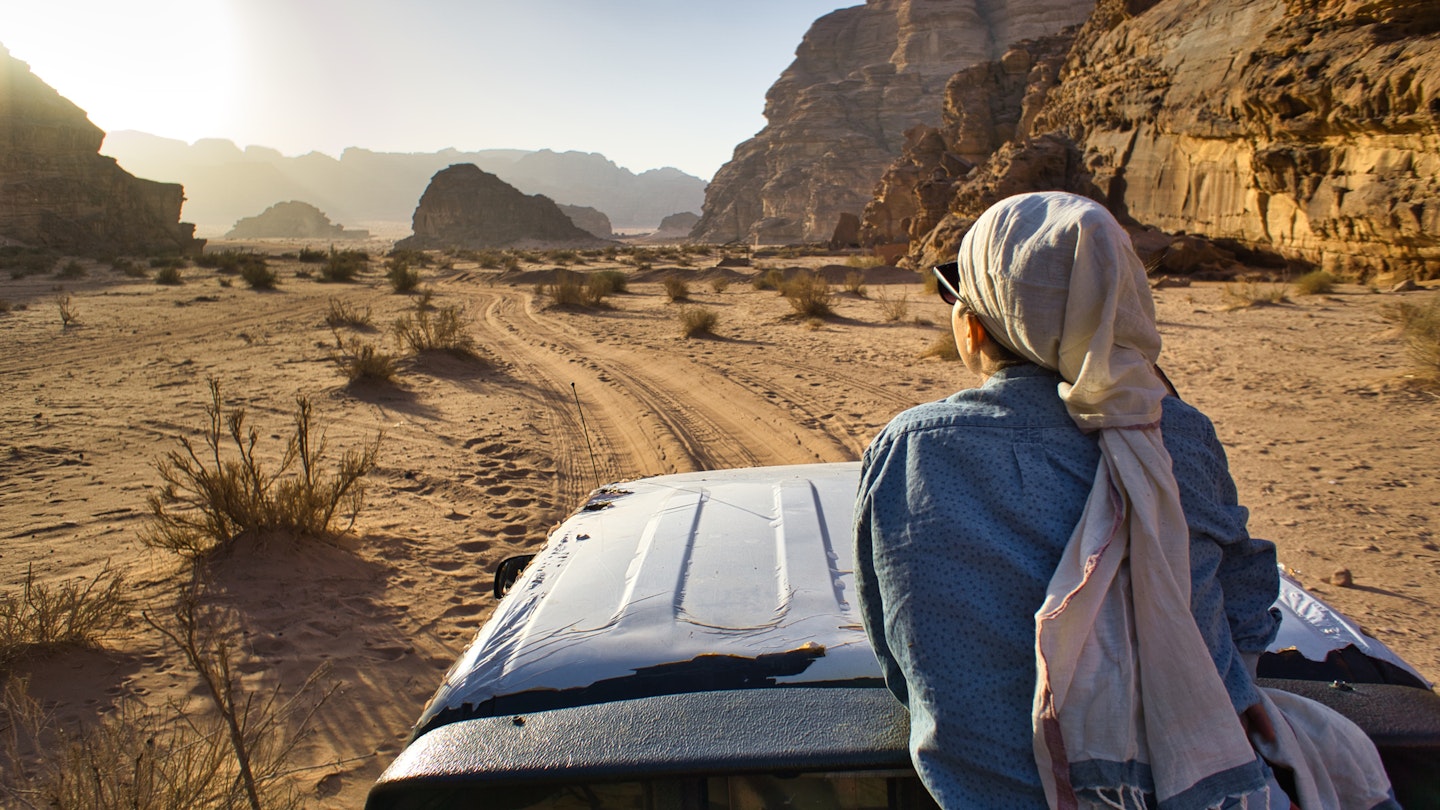 A woman tourist sitting on a car and admiring the sun light in an opening between two hills in the desert, during day time, in Wadi Rum, Jordan.