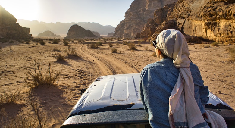 A woman tourist sitting on a car and admiring the sun light in an opening between two hills in the desert, during day time, in Wadi Rum, Jordan.