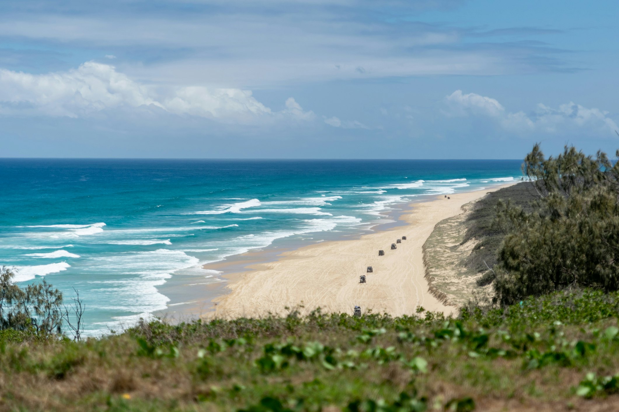 A vast sandy beah stretches into the distance with a row of 4WD vehicles driving along it