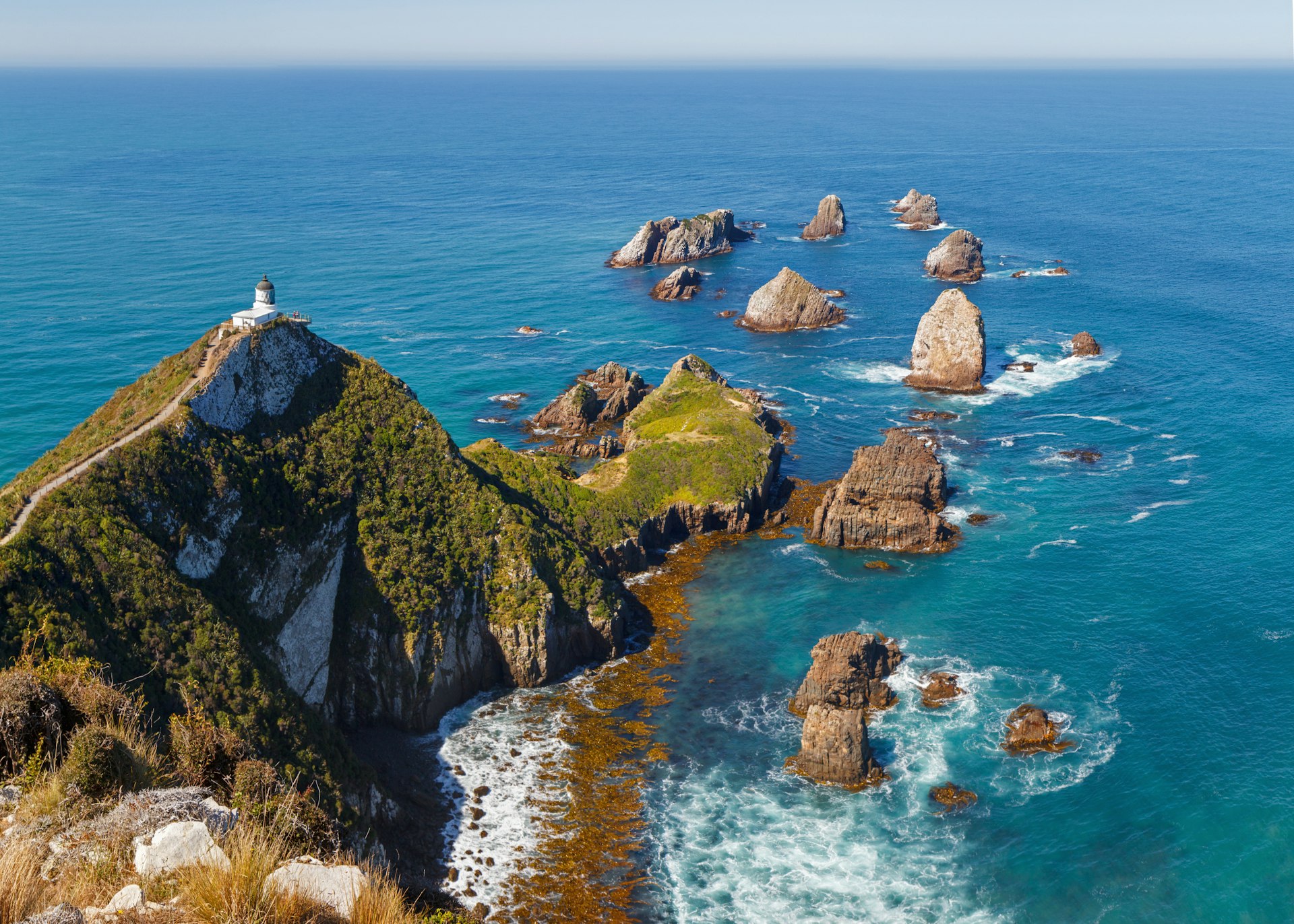 Nugget Point Lighthouse, New Zealand, sitting atop a forested coastal peak, with several rocky islands strung out into the sea beyond.