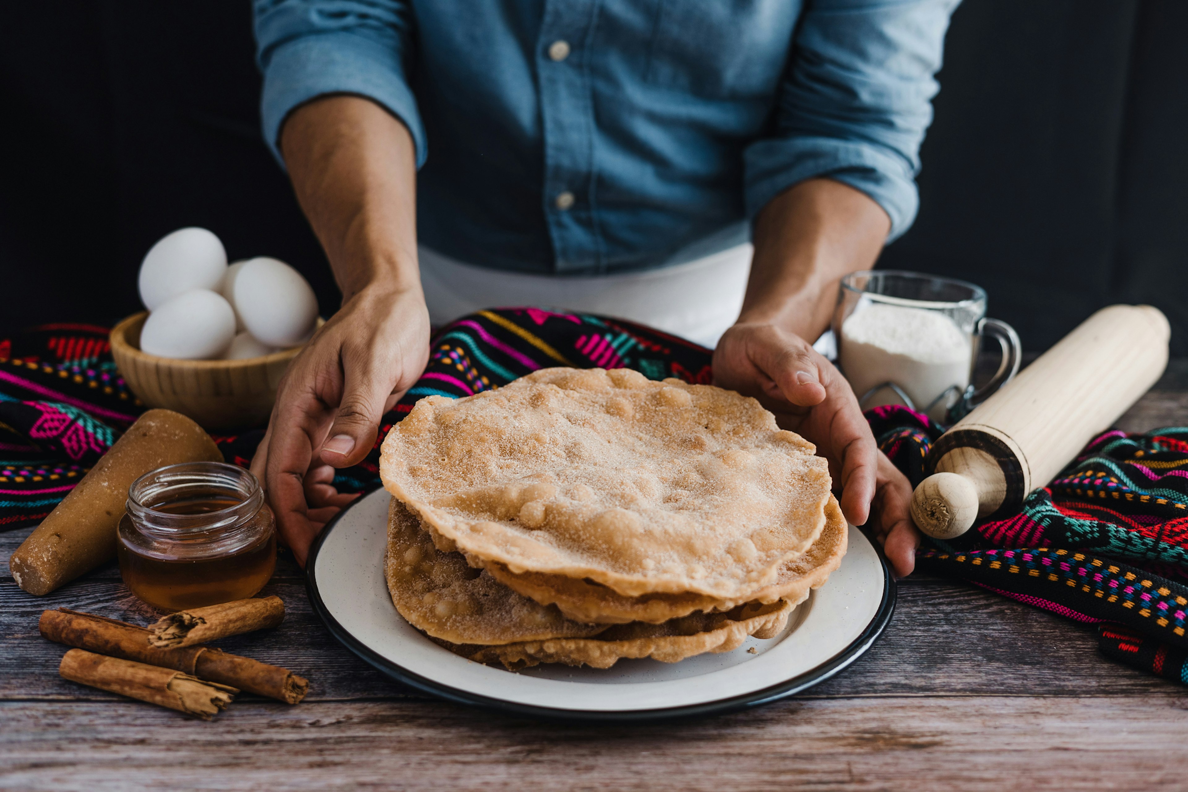 A man presents the flat, disc-like Buñuelos dessert on a table with Mexican patterning