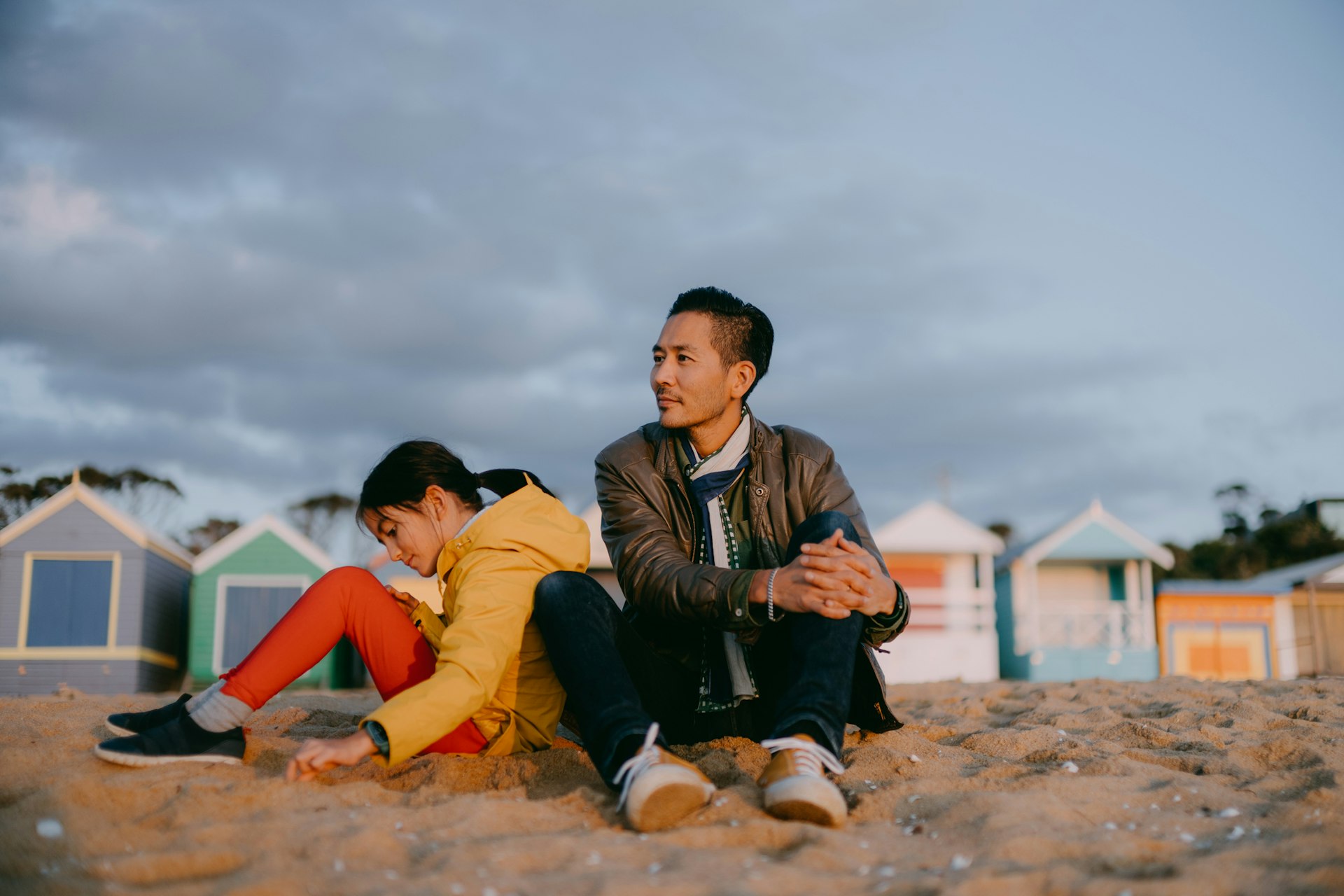 A father and daughter sitting on the beach together in Melbourne with colorful beach huts in the background