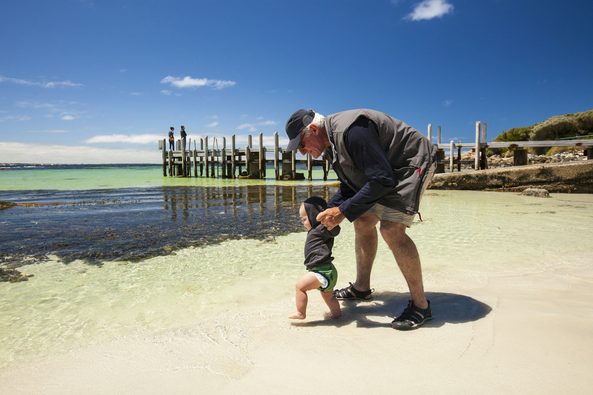 A grandad holds the hands of a toddler on the beach, who is heading straight into the ocean
