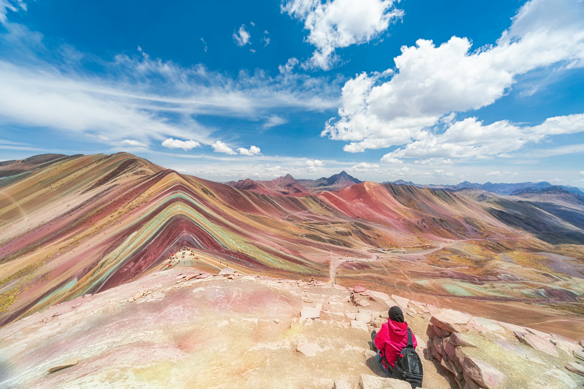 Female tourist sits in the foreground facing the colorful striated peaks of Vinicunca, or Rainbow Mountain.
