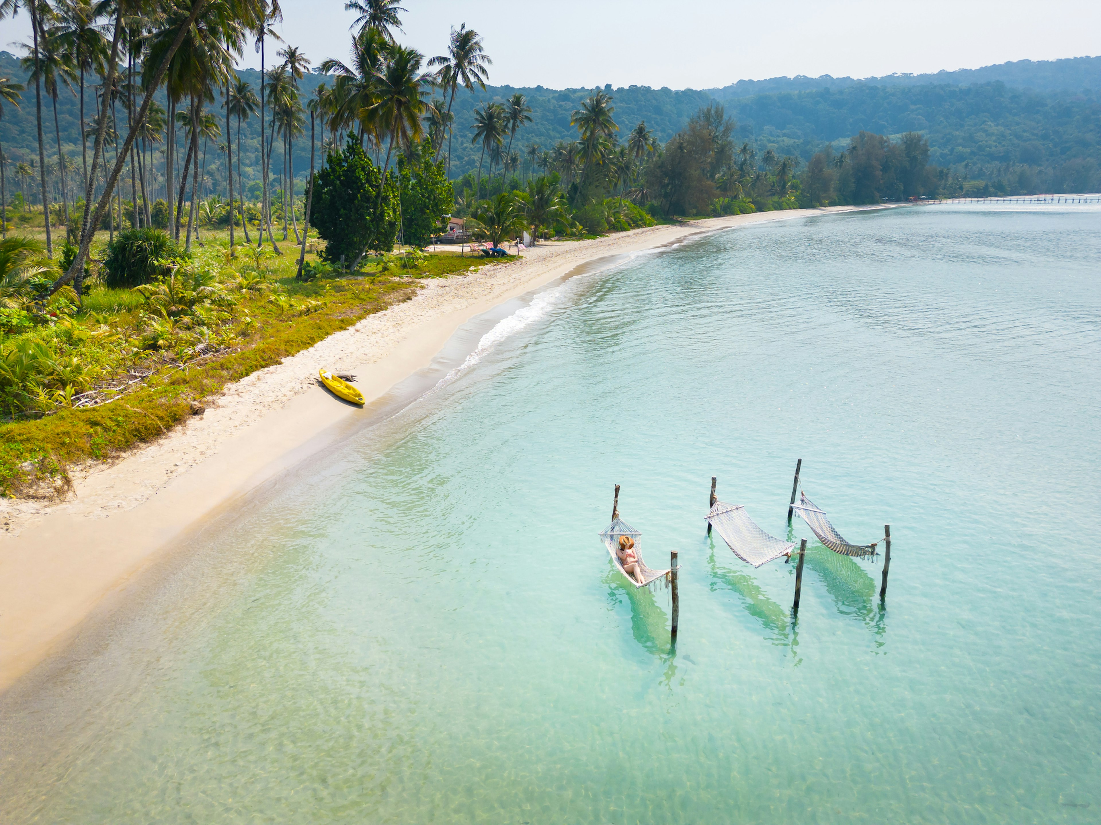 A woman lies in a hammock that's been strung on a frame out at sea. An empty sandy beach backed by palms stretches into the distance