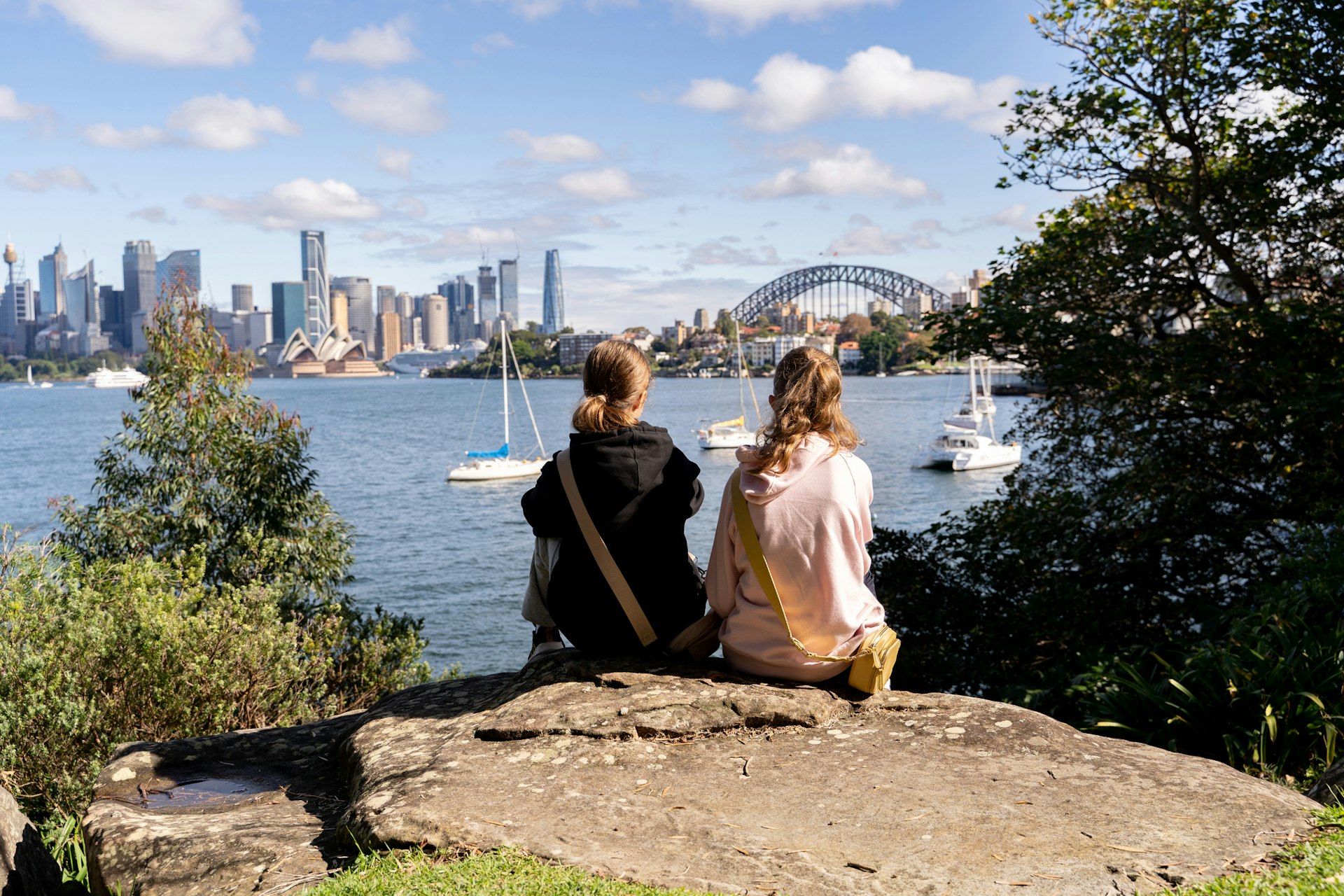 Two girls sit on a rock and look out over an iconic harbor, with a white opera house and a large bridge