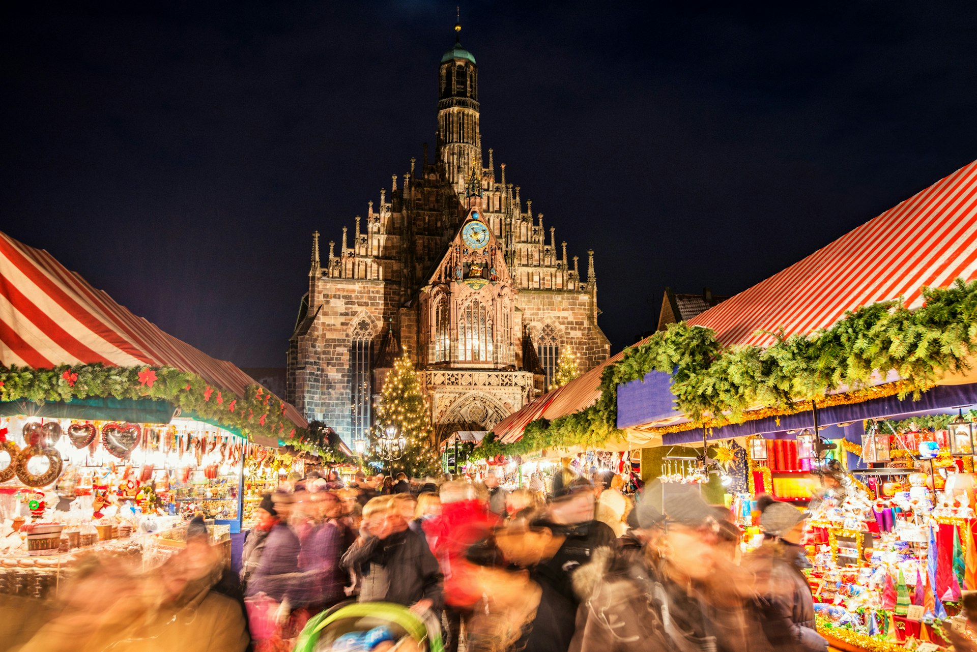 A huge crowd of people moving over Nuremberg's world-famous Christmas market (Nuremberg Christkindlesmarkt) at night, passing colorful illuminated Christmas decorations and food stalls.