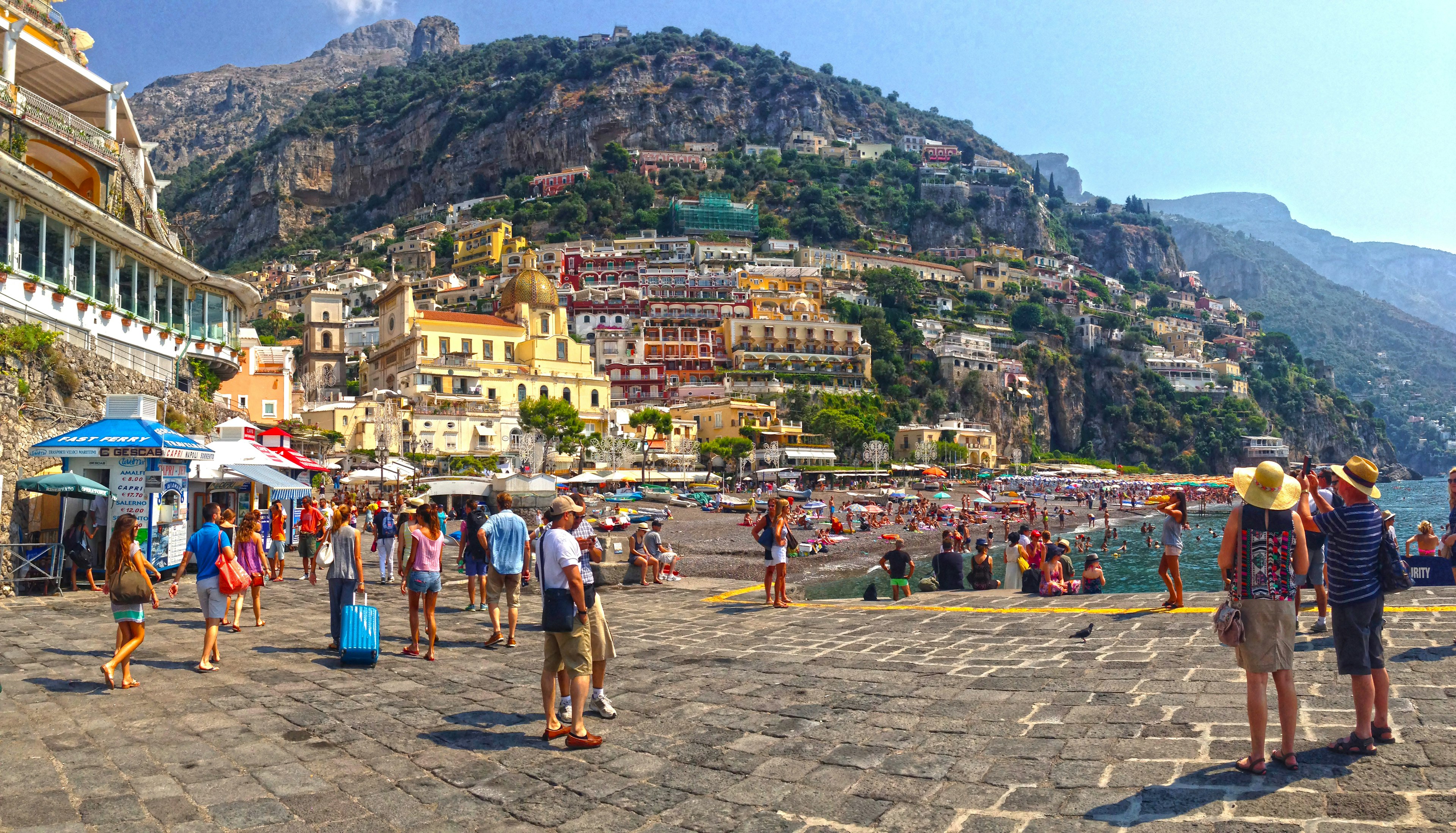 Tourists wander around the beachfront of a town with buildings built into the cliff