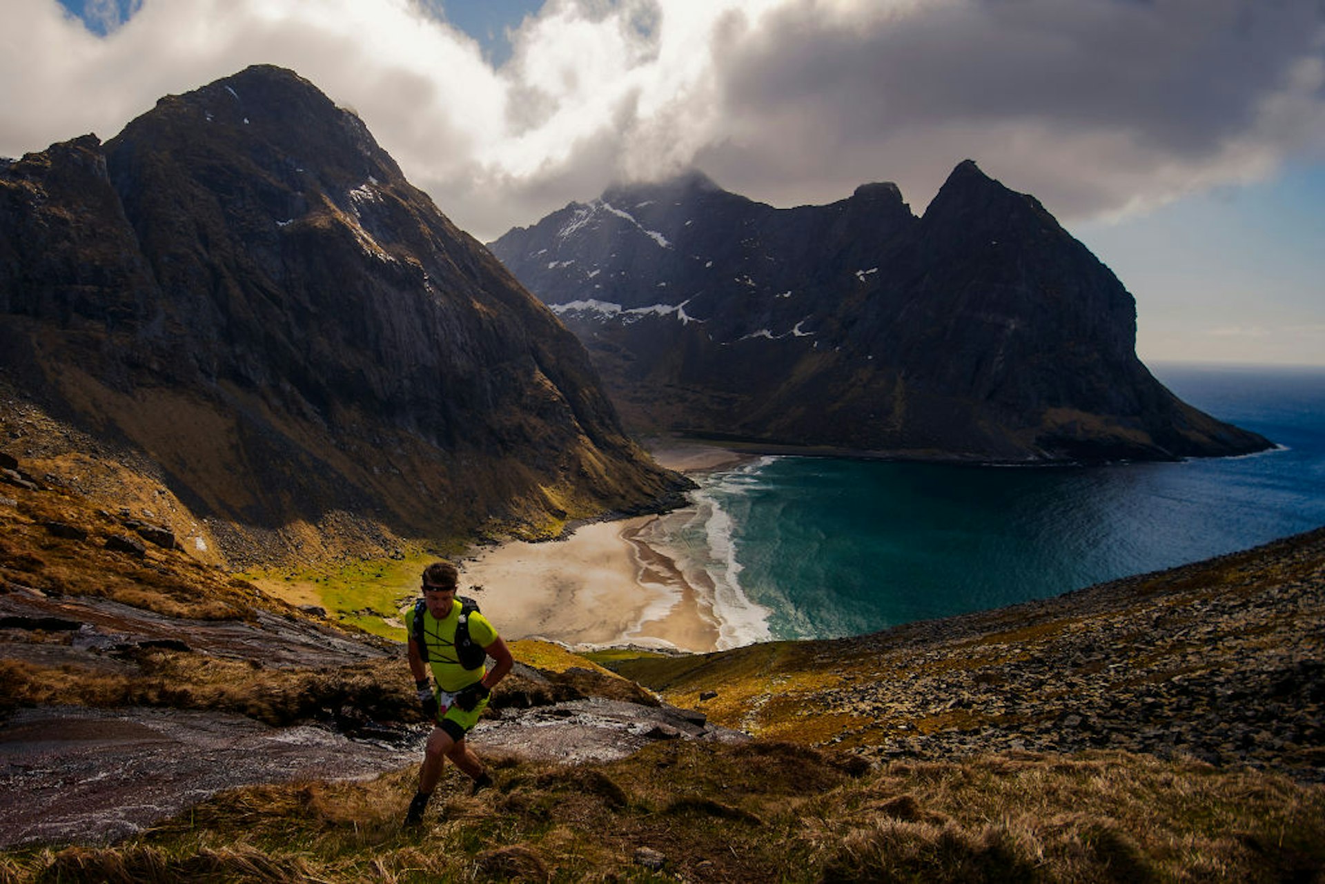 A hiker in bright yellow clothes climbs up the mountainside with Kvalvika Beach, Lofoten Islands, in the background.