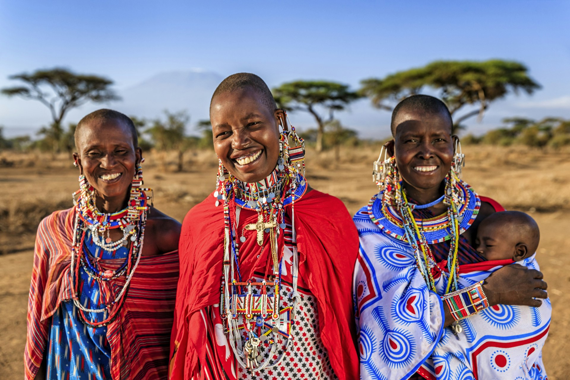 Three smiling Maasai women in colorful prints, one is holding a small baby