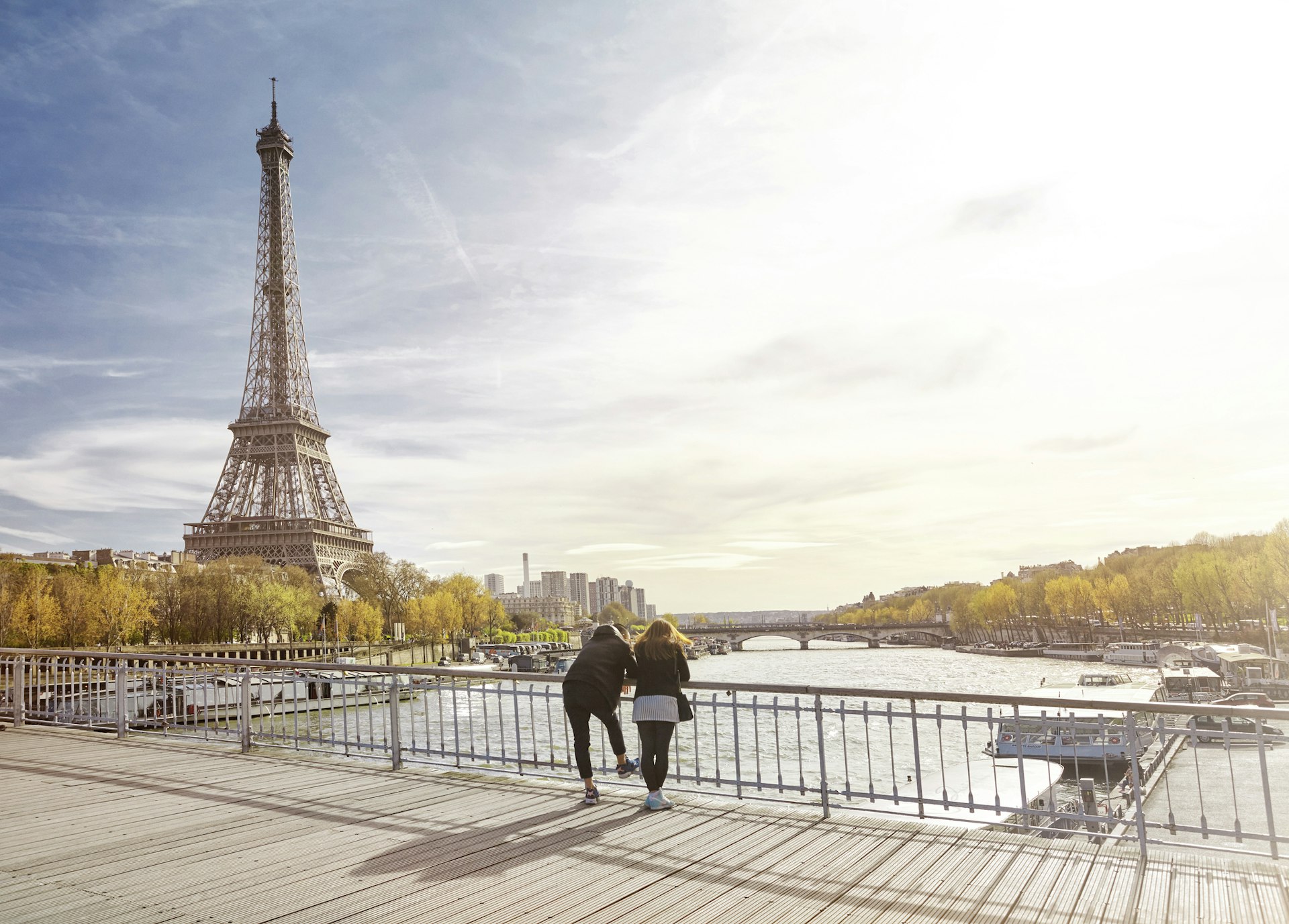 Two people lean on the edge of a bridge looking out over a river. The iconic Eiffel Tower dominates the skyline
