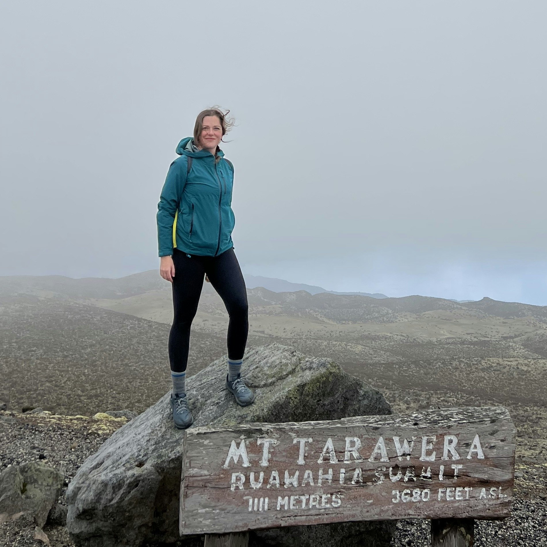 Jessica Lockhart in New Zealand, standing on a rock behind a wooden sign marking the summit of Mt Tarawera