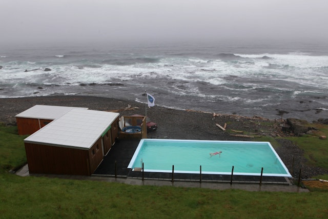 A woman swims in a rectangular pool heated by hot springs next to a black volcanic beach with the sea raging in the distance