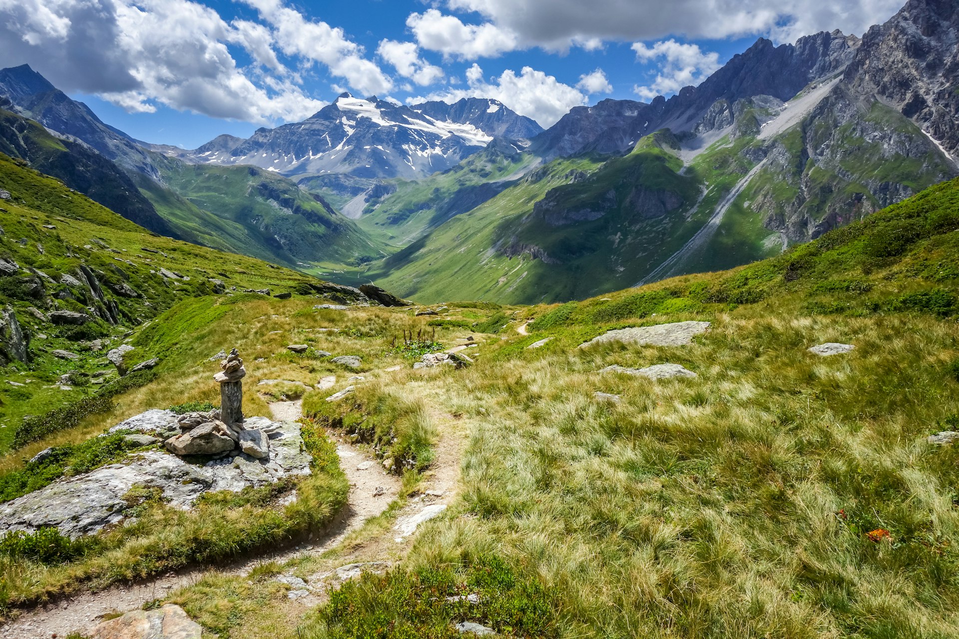 A rugged trail winds through a rocky, grassy alpine landscape, with snow-dusted mountains in the distance