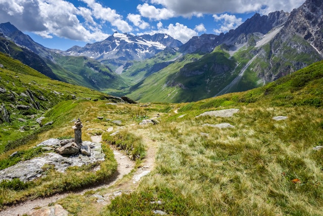 A rugged trail winds through a rocky, grassy alpine landscape, with snow-dusted mountains in the distance
