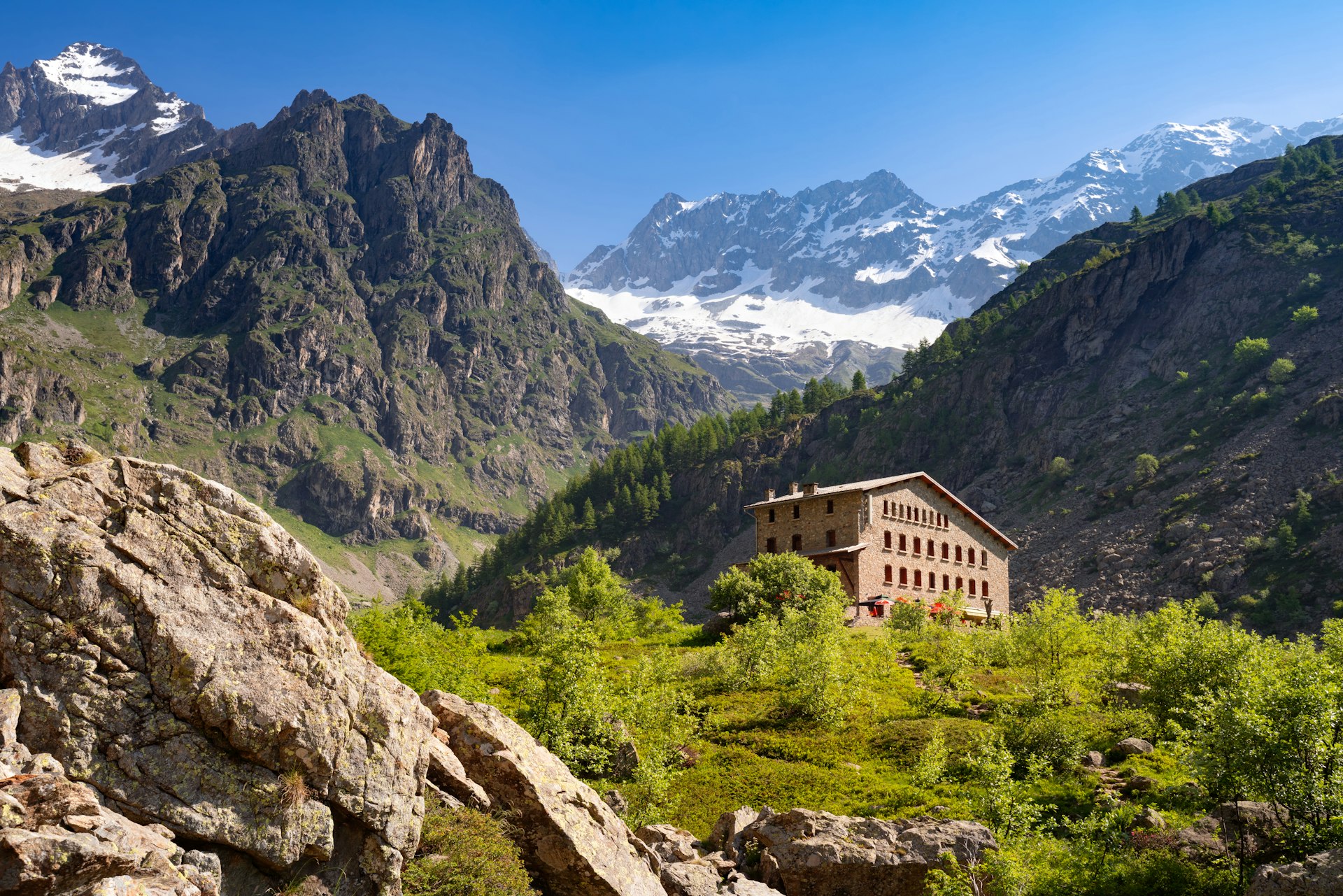 Um edifício de pedra como refúgio nas montanhas, escondido nas colinas, com uma montanha coberta de neve erguendo-se acima 