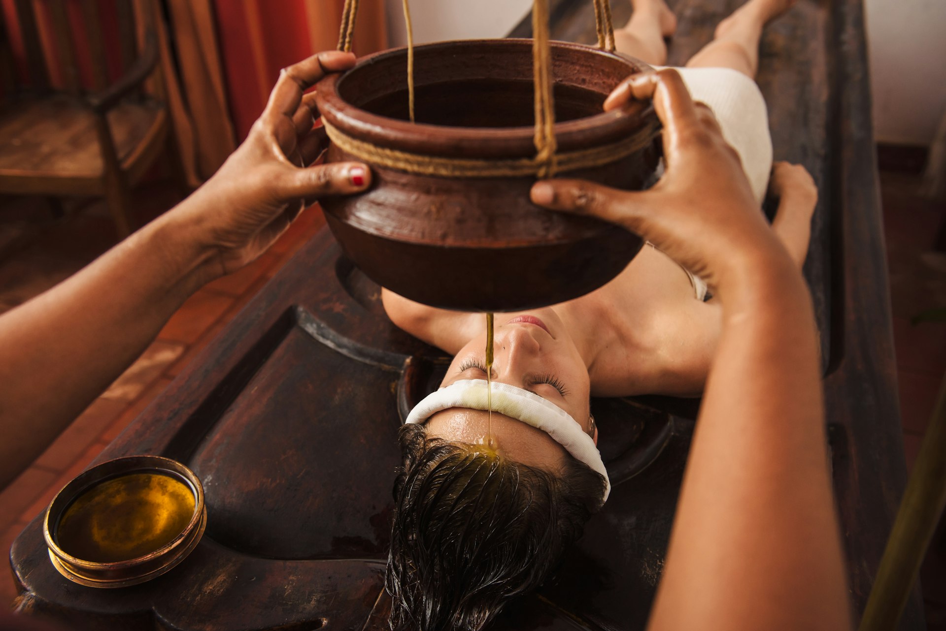 A woman lies on her back in a spa, as a honey-coloured liquid is poured on her forehead as part of an Ayurvedic wellness treatment