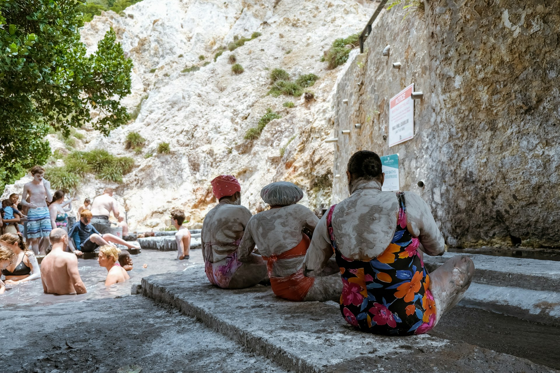 Three women in swimwear sit on the edge of a mud bath completely covered in a gray coating of sulphur-rich mud