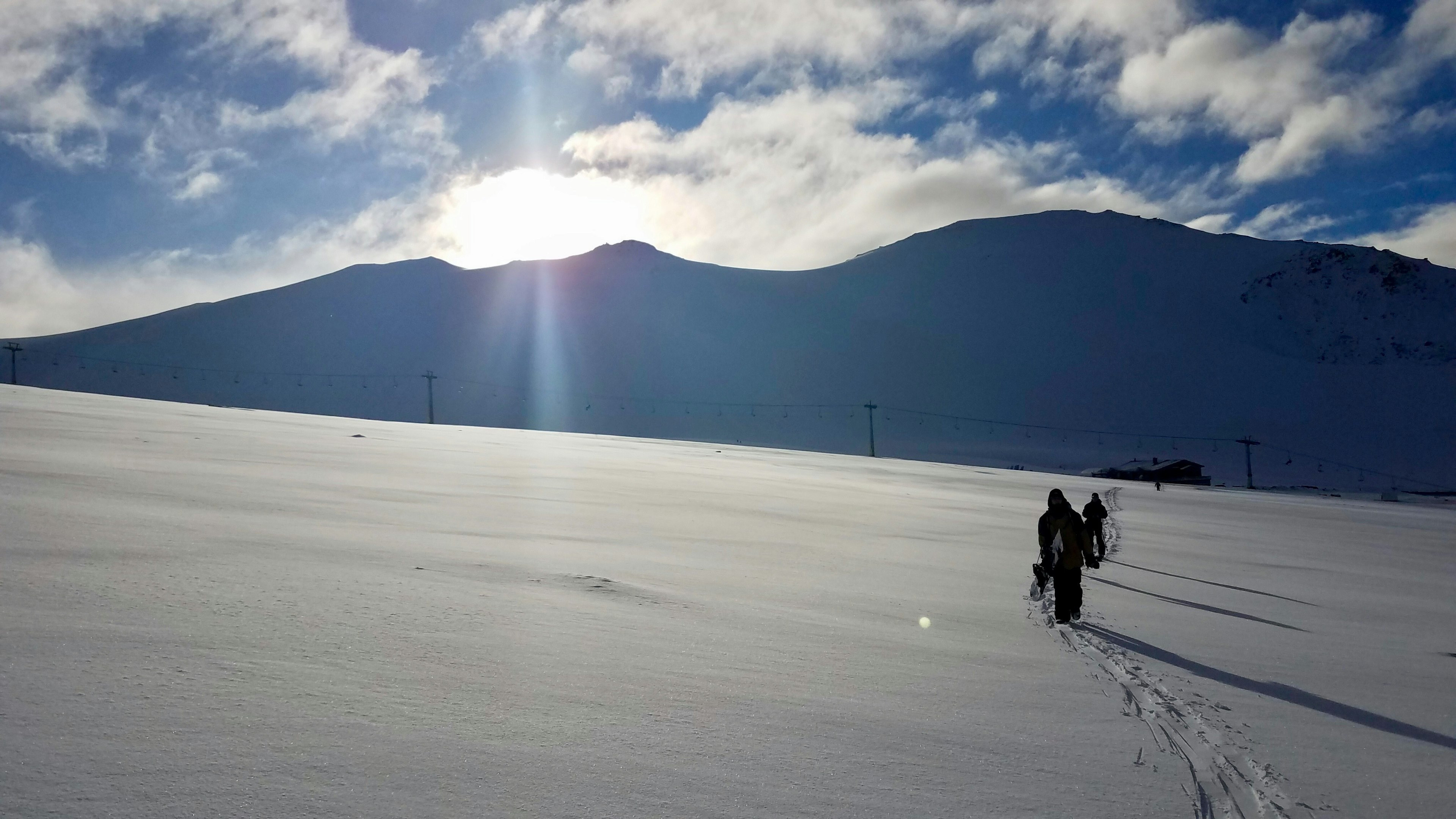 Snowboarders tramp across a vast, flat expanse of pristine snow, carrying their snowboards