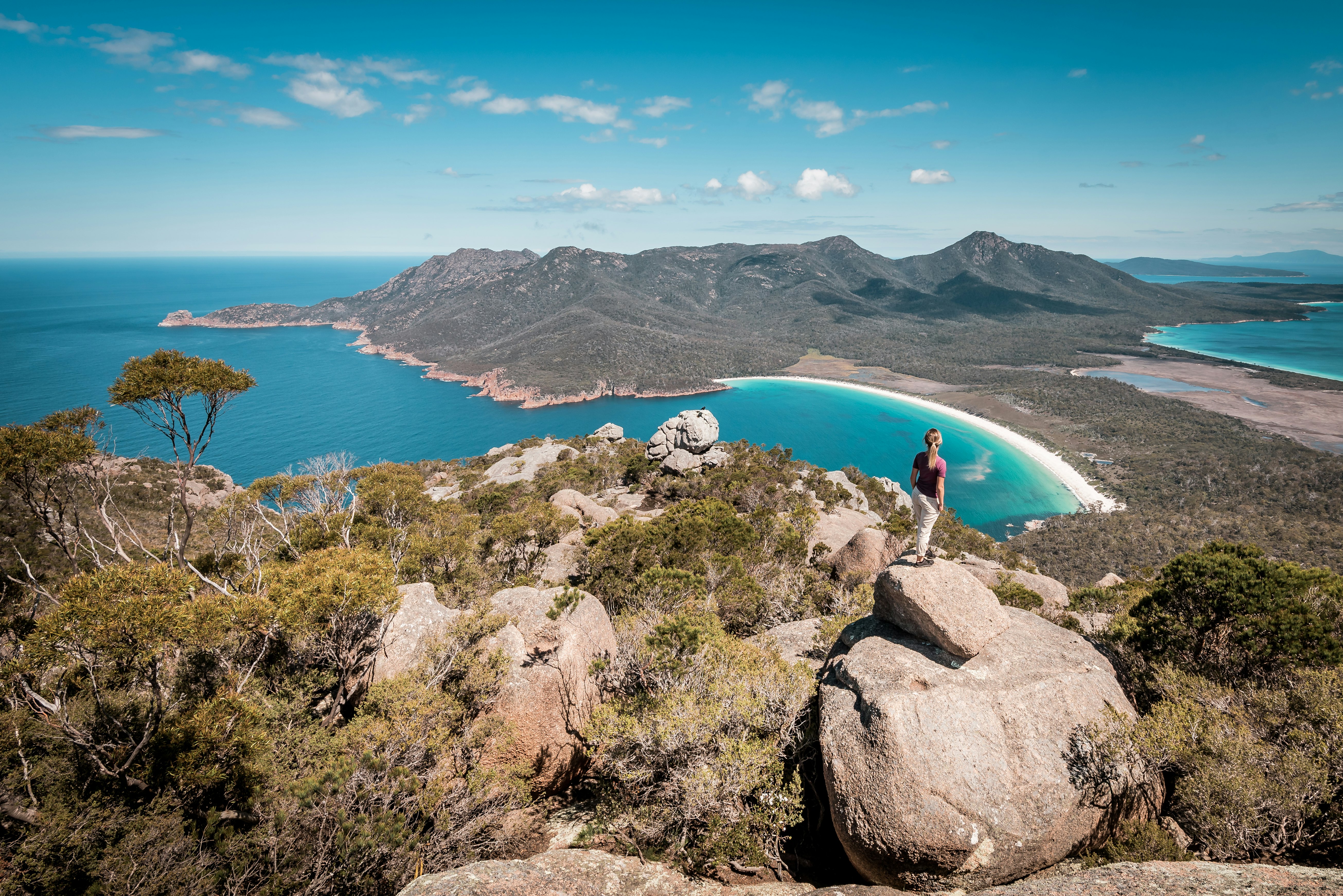 A hikers stands at a viewpoint looking down to a beautiful curve of beach