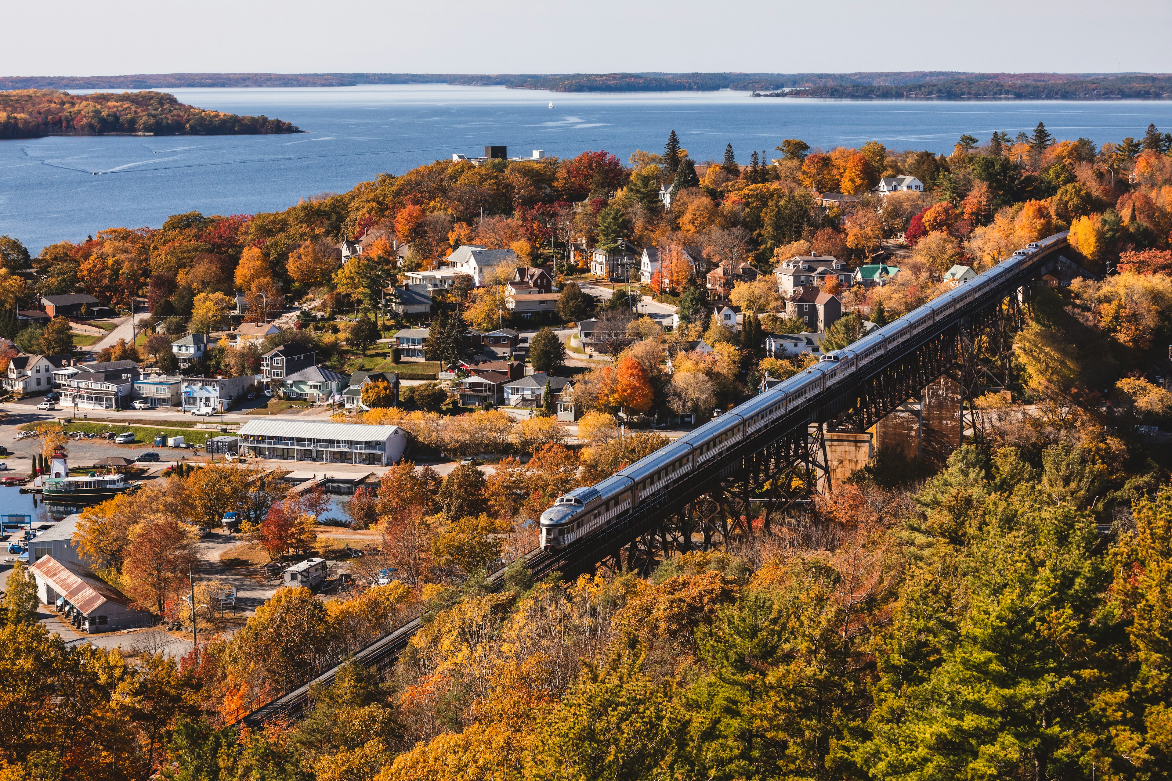 An aerial view of a train crossing a trestle by a charming waterfront town filled with trees in foliage