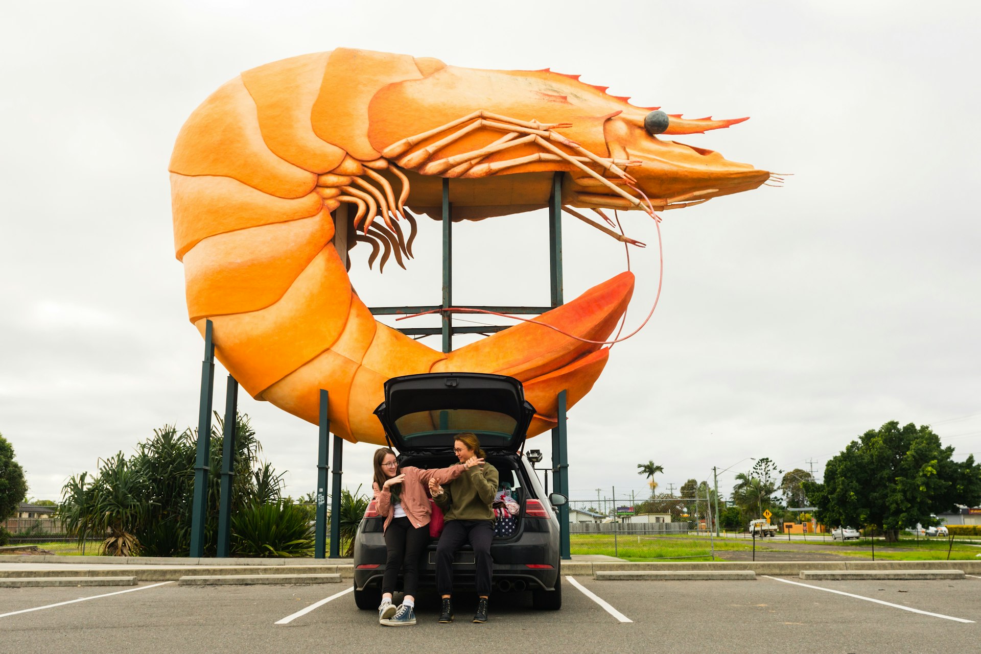 Two kids lean on the trunk of a car parked underneath a huge sculpture of a prawn/shrimp