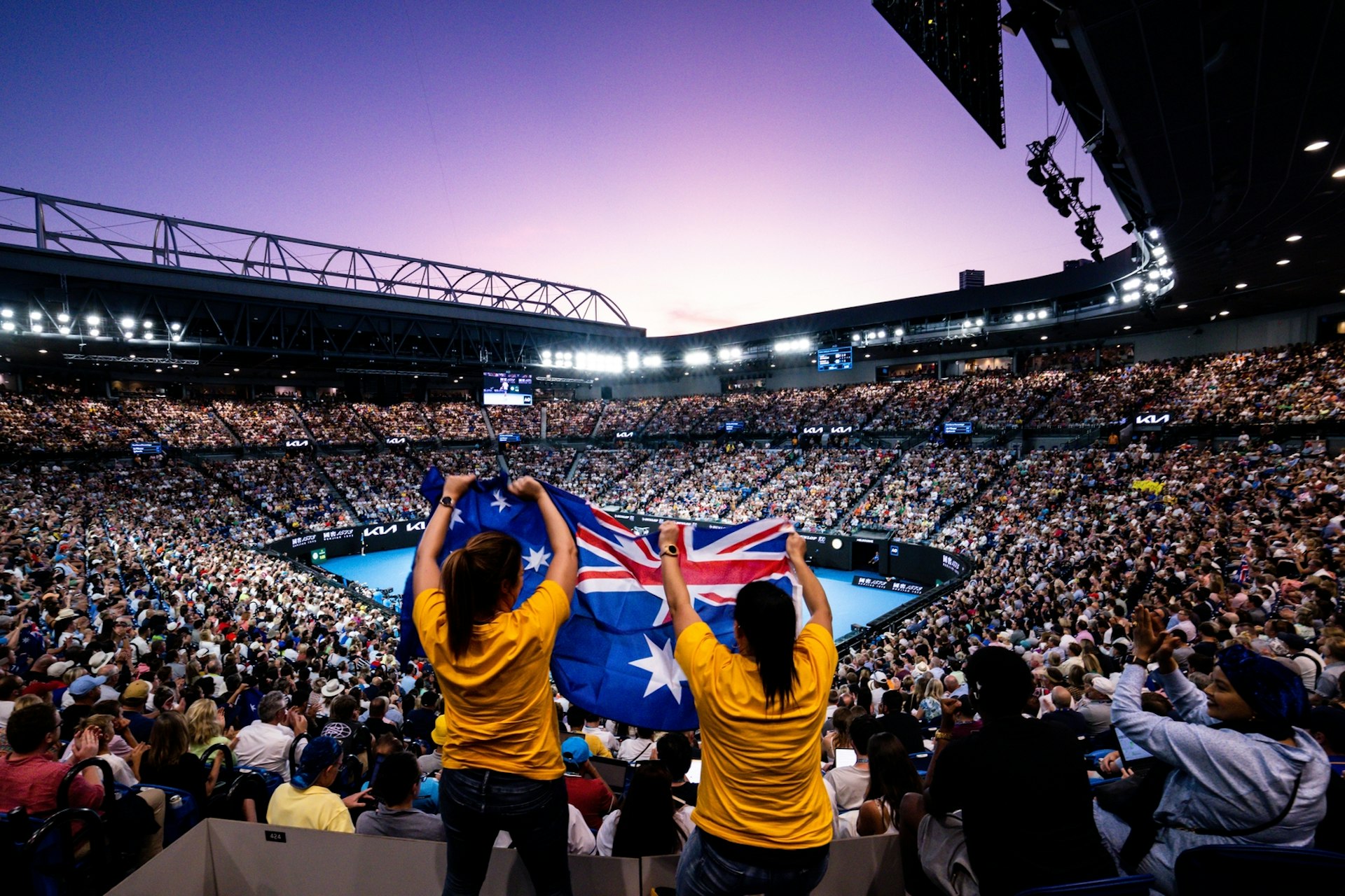 Two people stand up among a crowd at a tennis tournament waving an Australian flag
