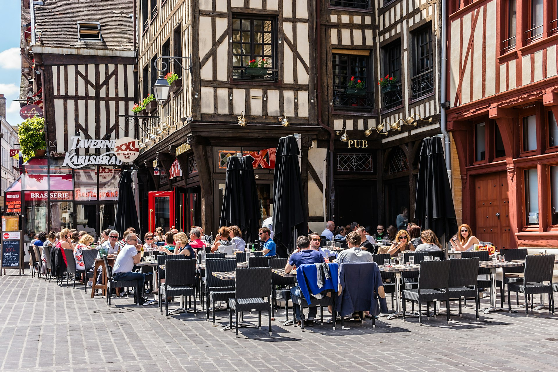 People sit at outside tables in a pedestrianized city square surrounded by historic half-timbered houses
