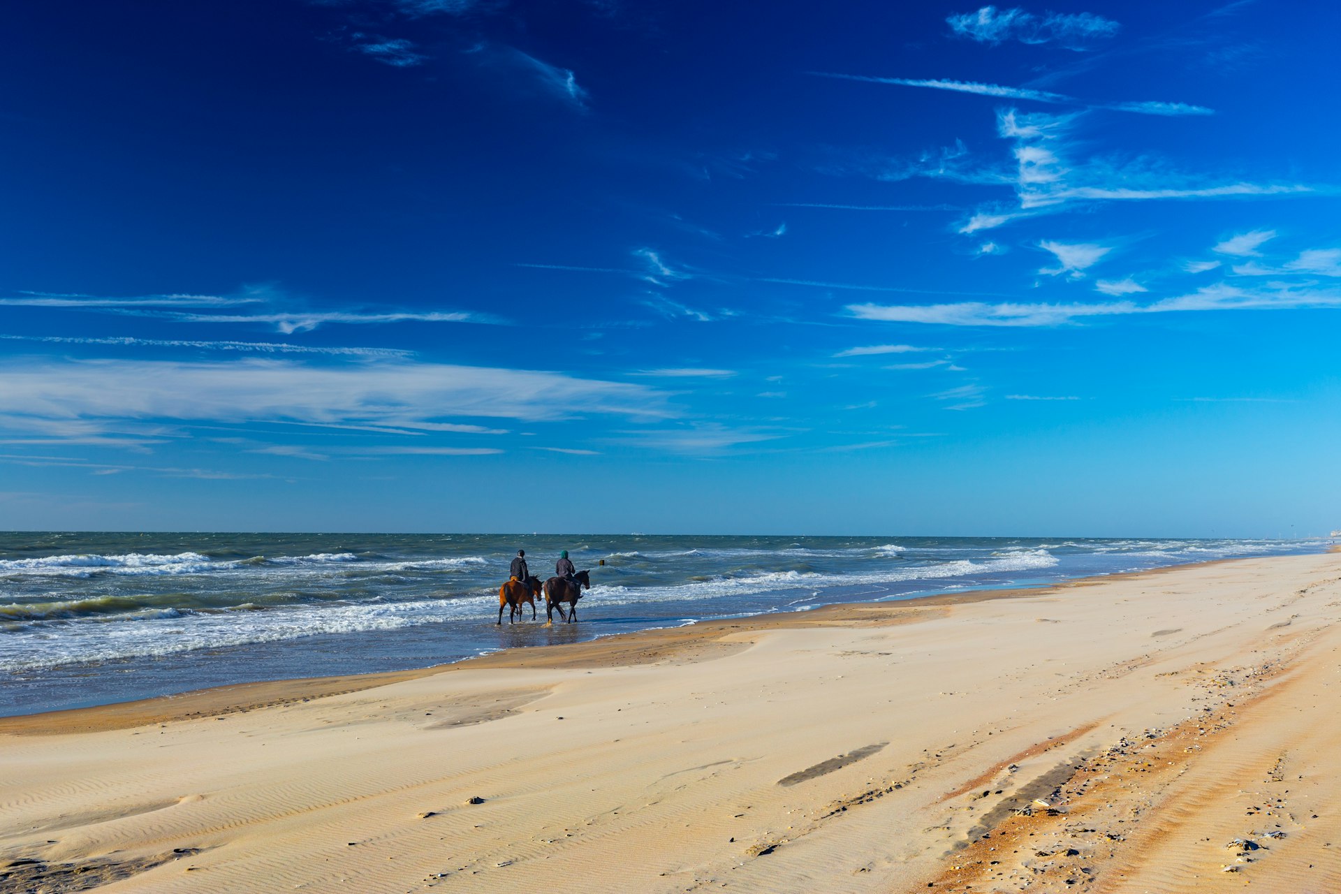 Two people on horseback ride along the beach on a sunny day