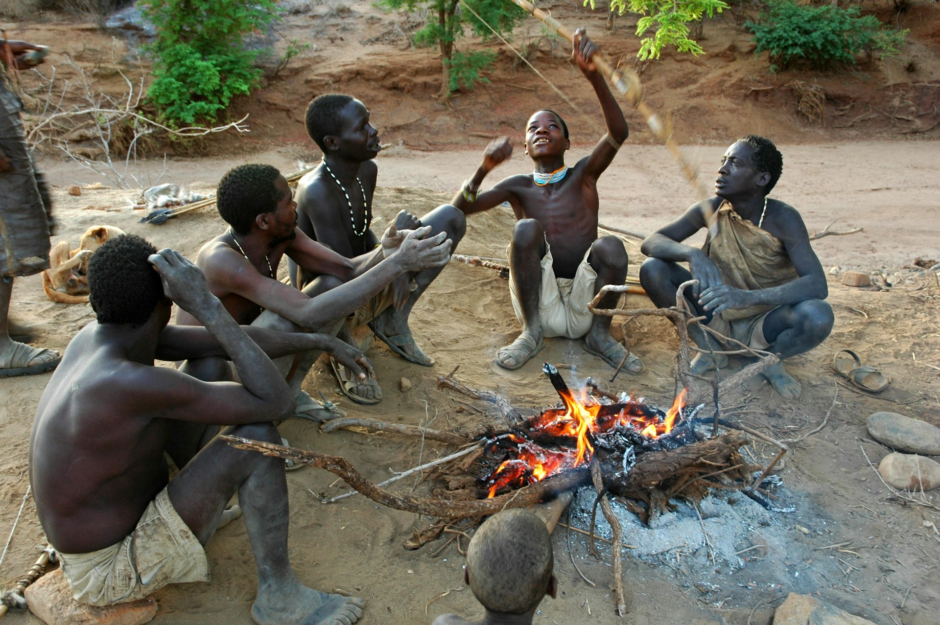 Group of Hadzabe tribesmen sit around fire with bow and arrow.