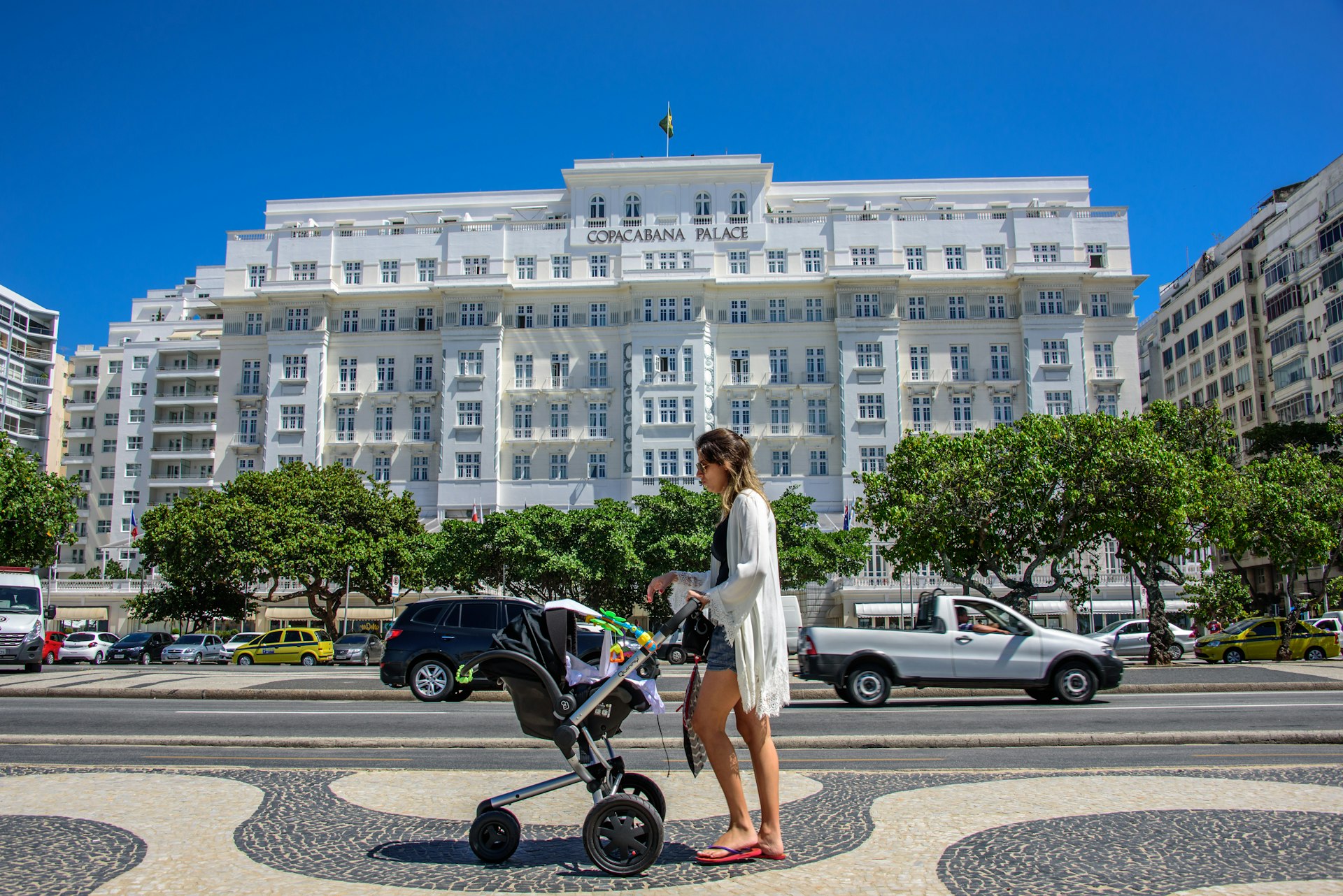 A woman wearing a white dress and flip flops walks along Avenida Atlantica with a black baby stroller, with Copacabana Palace in the background