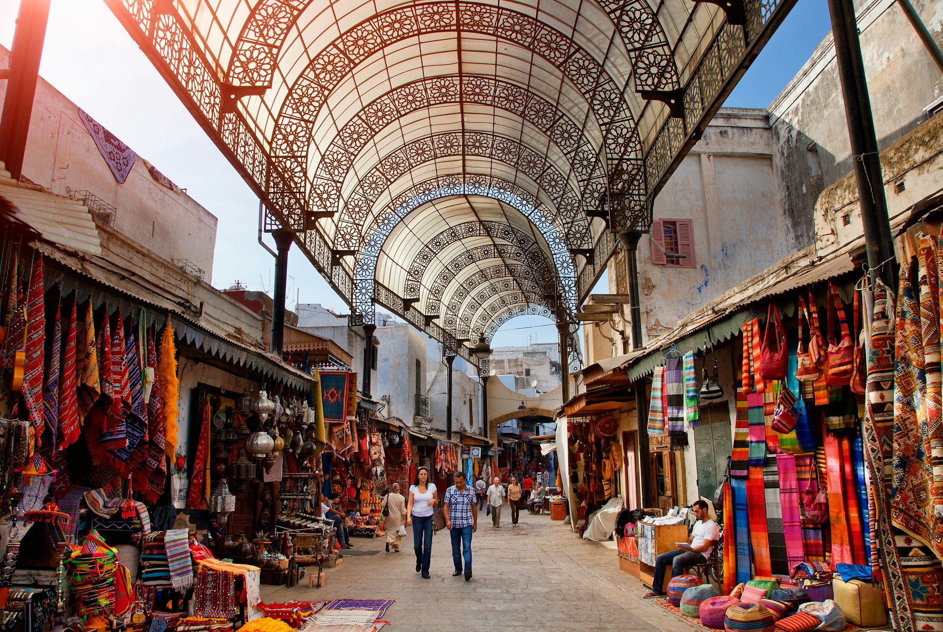 Travelers shop in a covered market, where stallholders sell colorful rugs and scarves 