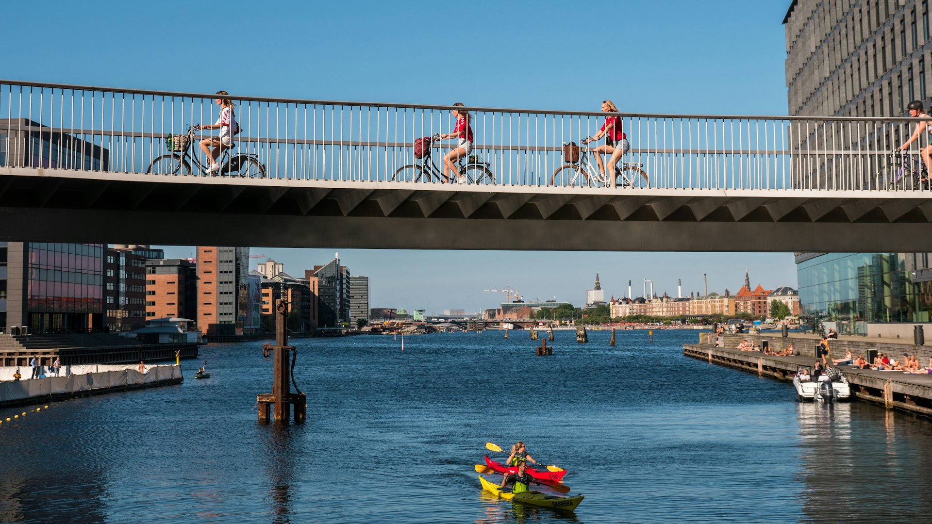 People cycling over a bridge while others kayak in the water below them in Copenhagen