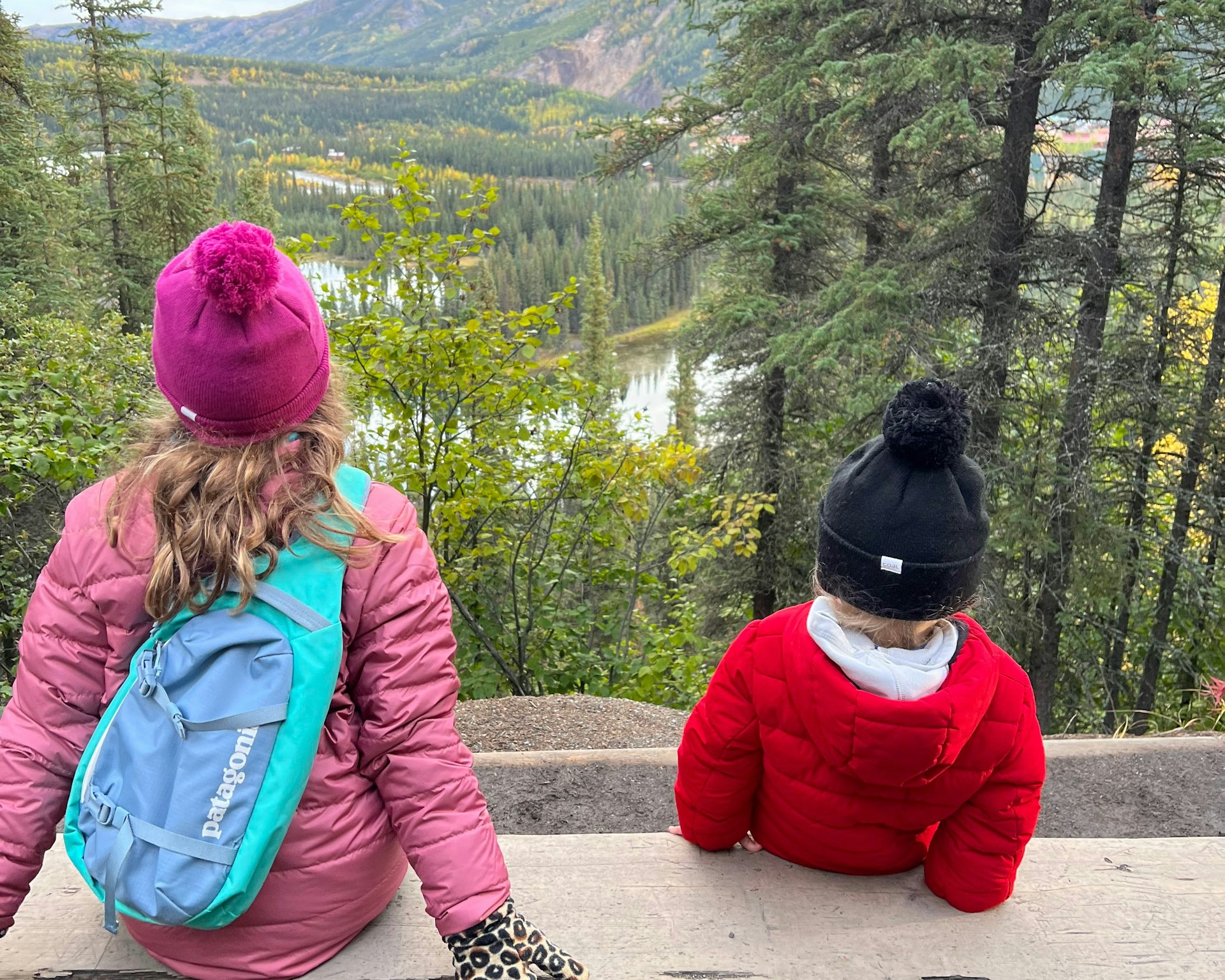 Two children in bobble hats look out across Beaver Pond in Alaska which is surrounded by evergreen trees