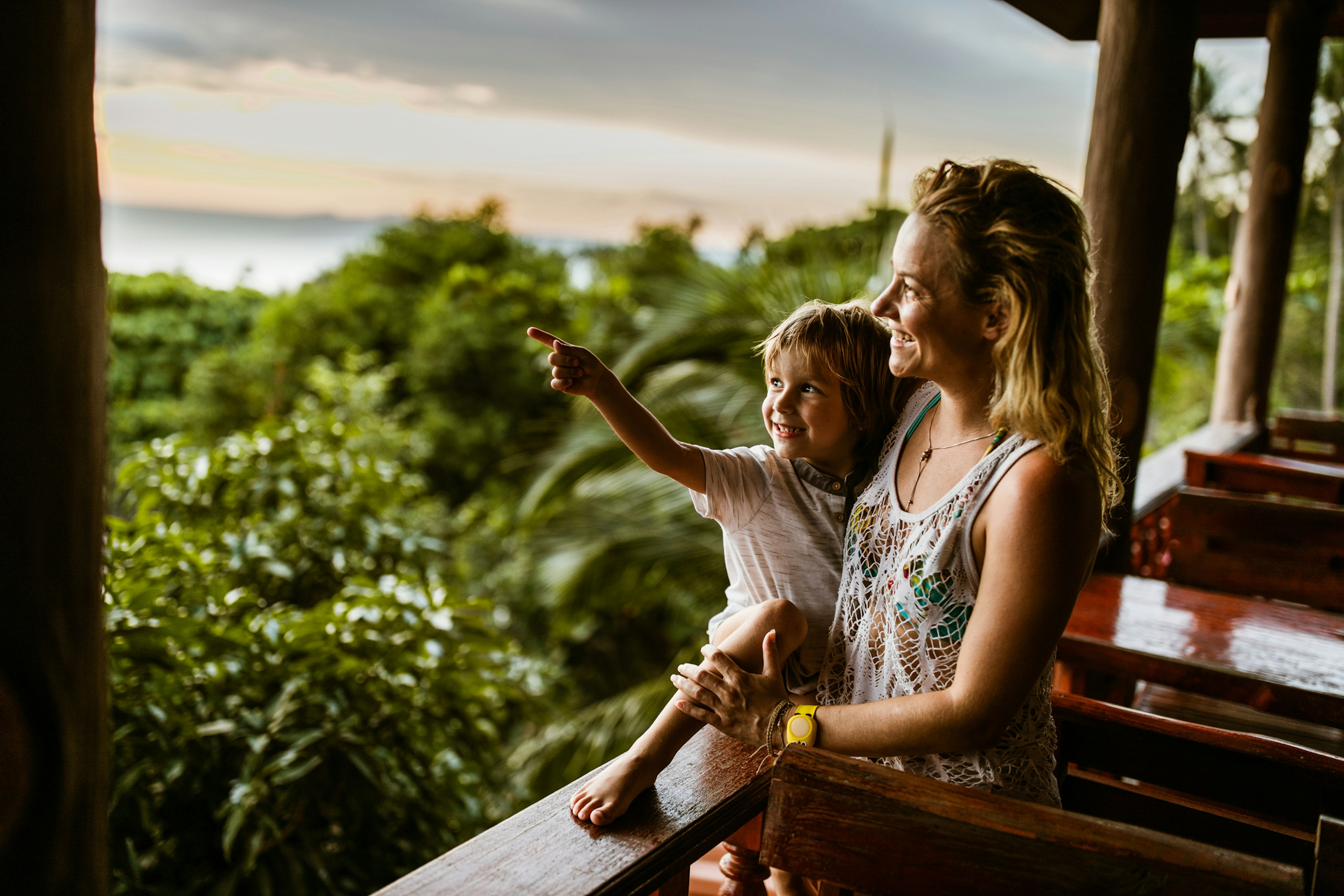 A small boy in his mother's arms smiles and points out towards the jungle-like environment near their balcony