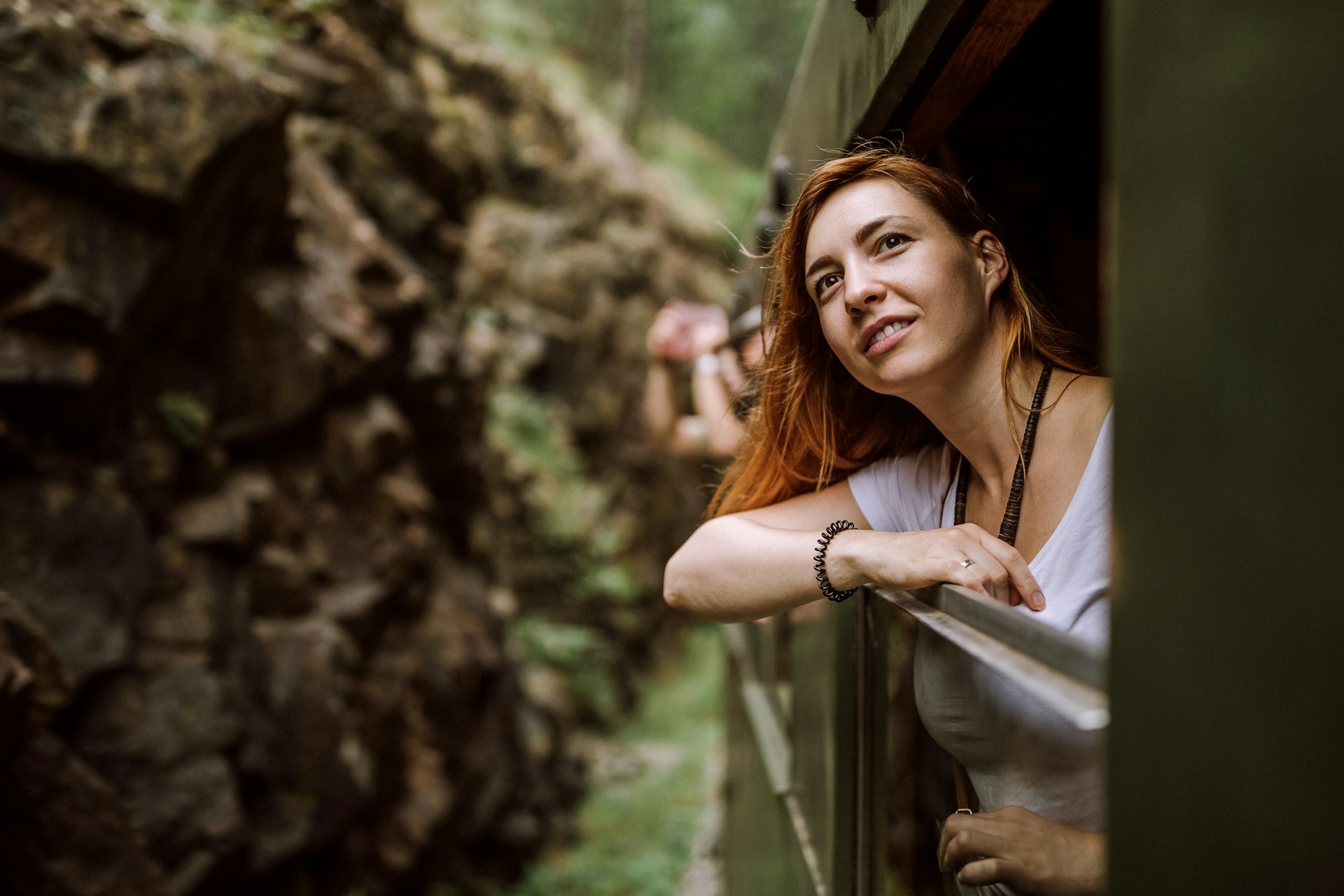 A woman is traveling on a train, leaning out of a window and looking out at the scenery