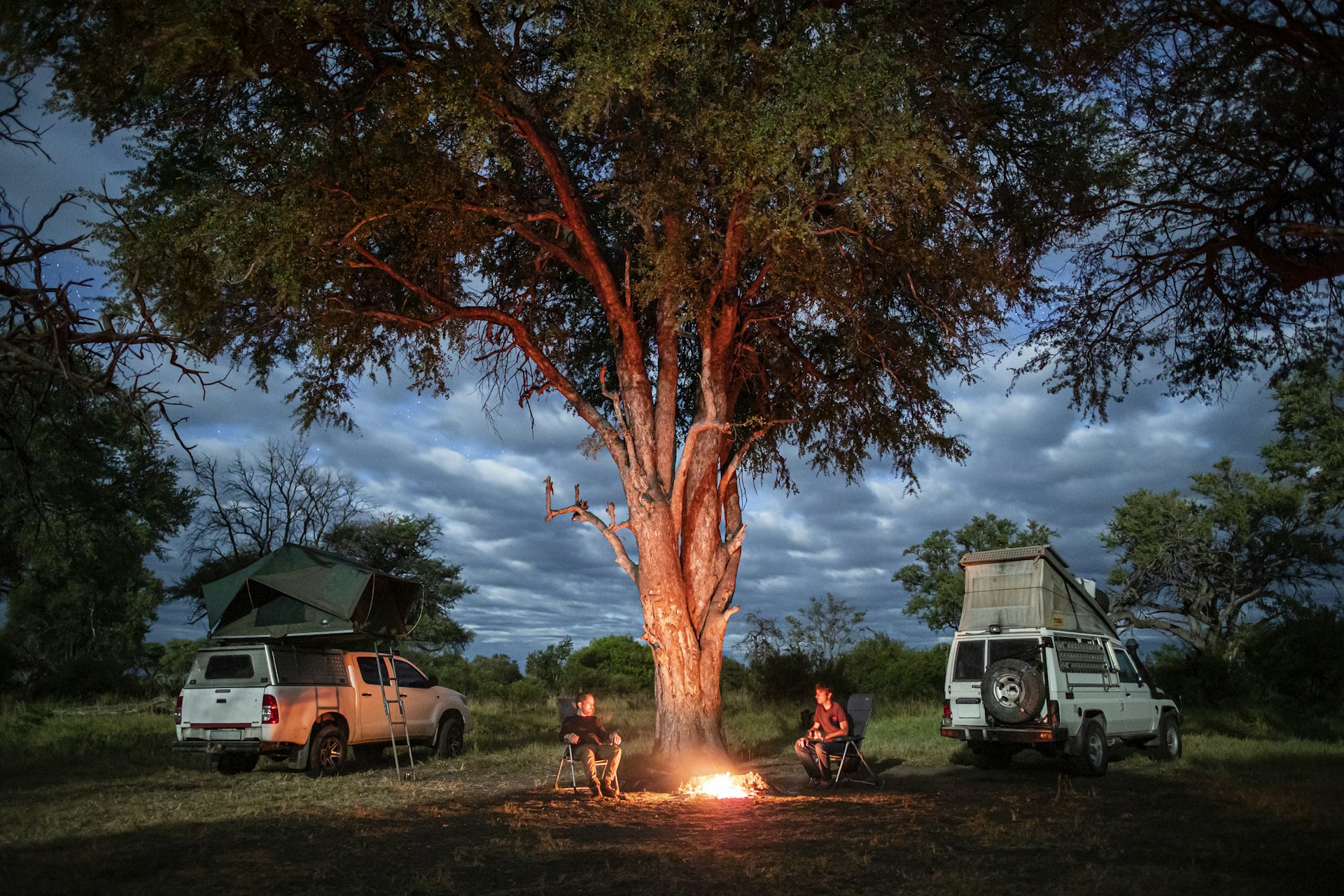 Two men sit near a fire beneath a tree in the African bush next to their camping 4WD cars which have the roof tents popped up