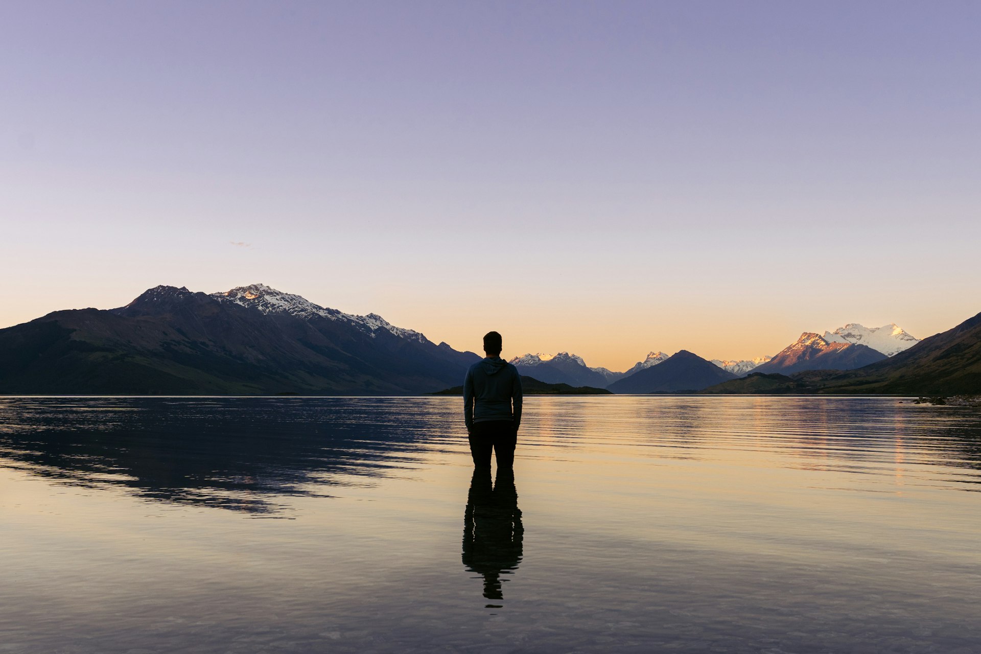 Man inside Lake Wakatipu looking at the amazing sunset behind snowy mountain peaks. New Zealand landscape