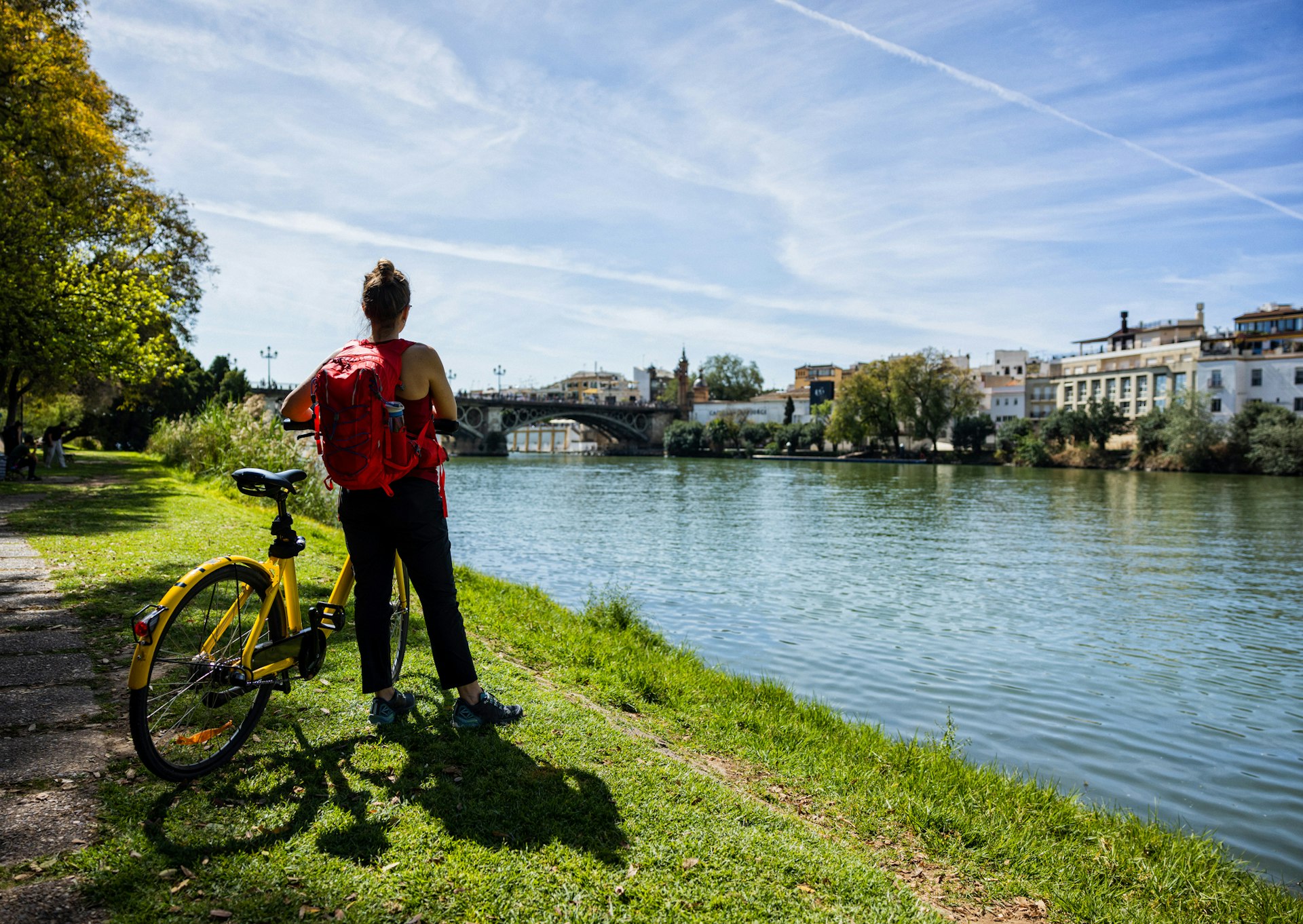 A tourist stands by a river beside her bike looking across at the city skyline on a sunny day 