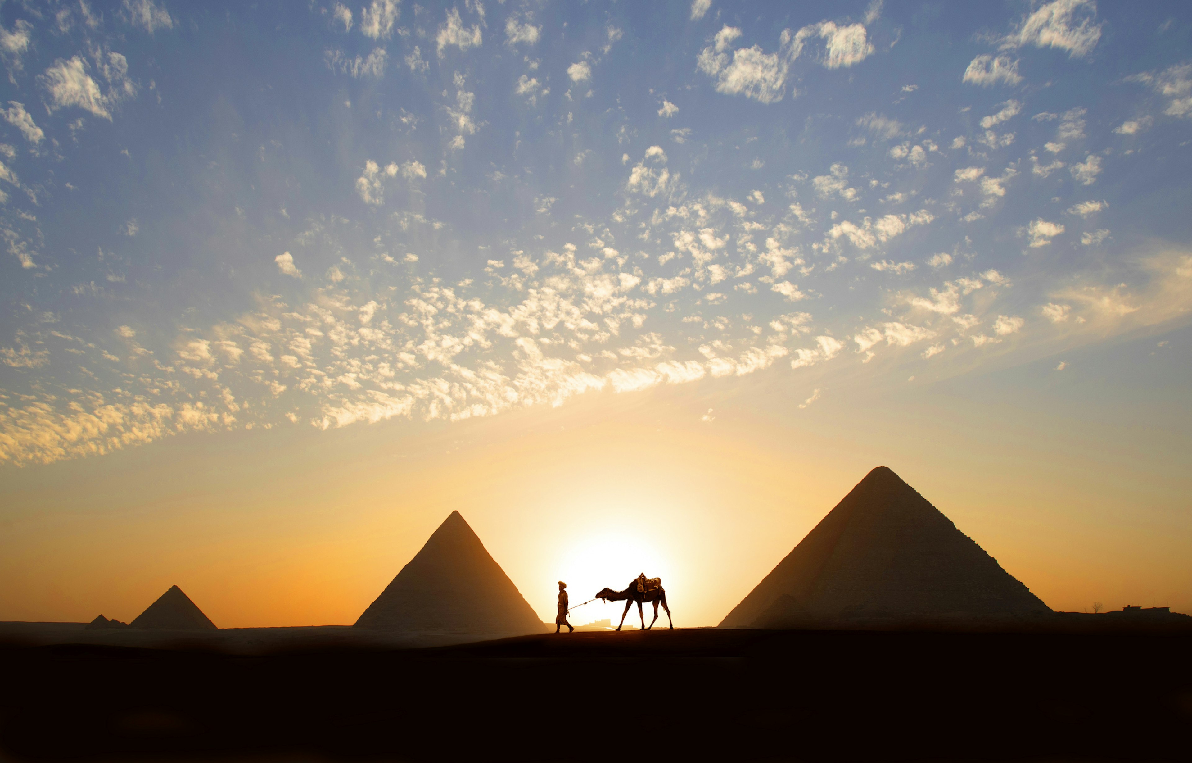 A single tour guide walking a camel at sunset by a huge stone pyramid in the desert