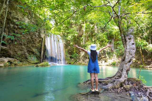 A teen stands facing a waterfall plunging into a blue pool with her arms in the air and her back to the camera