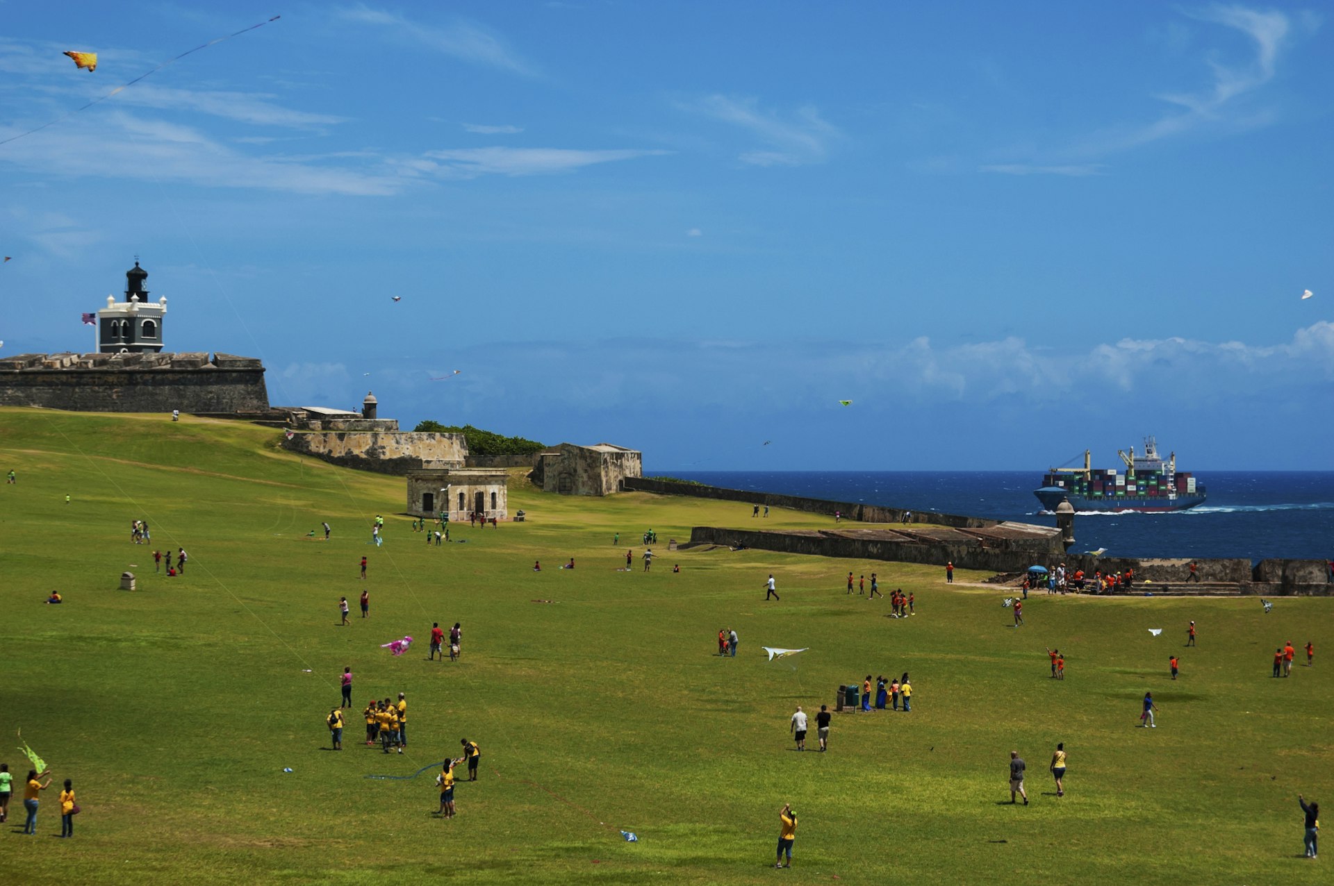 Children and parents are playing with kites in front of a seaside fortress 
