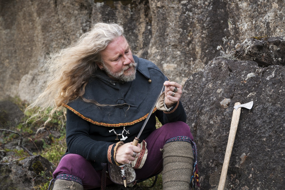 Blond Viking male reenactor in full warrior armour and battle gear is sitting on a rock cleaning his knife in the historic location where Vikings assembled annually to recite and discuss laws, Thingvellir National Park, Pingvellir, Iceland